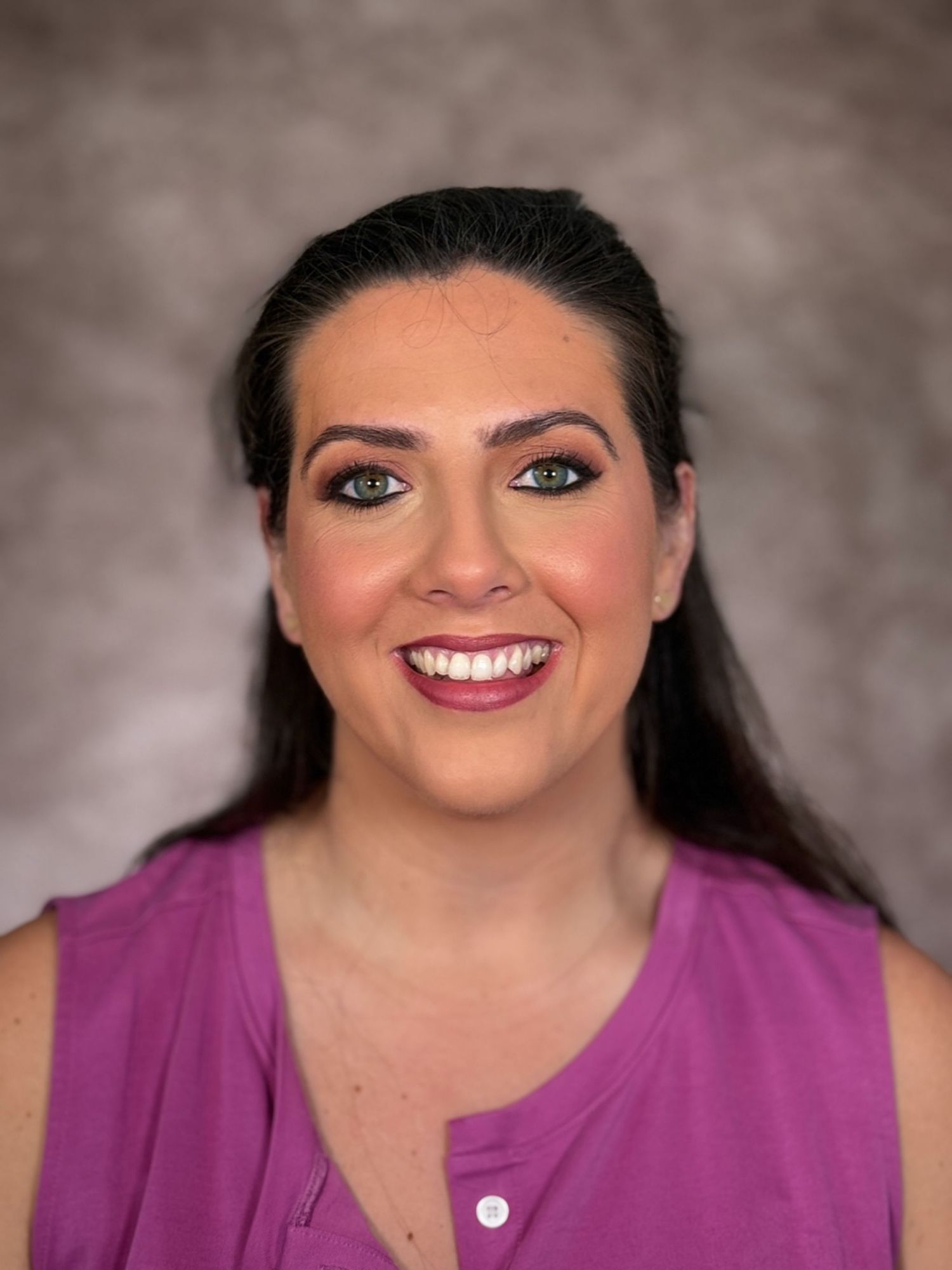 Professional headshot of a smiling woman with dark hair, wearing a purple sleeveless top against a gray background.