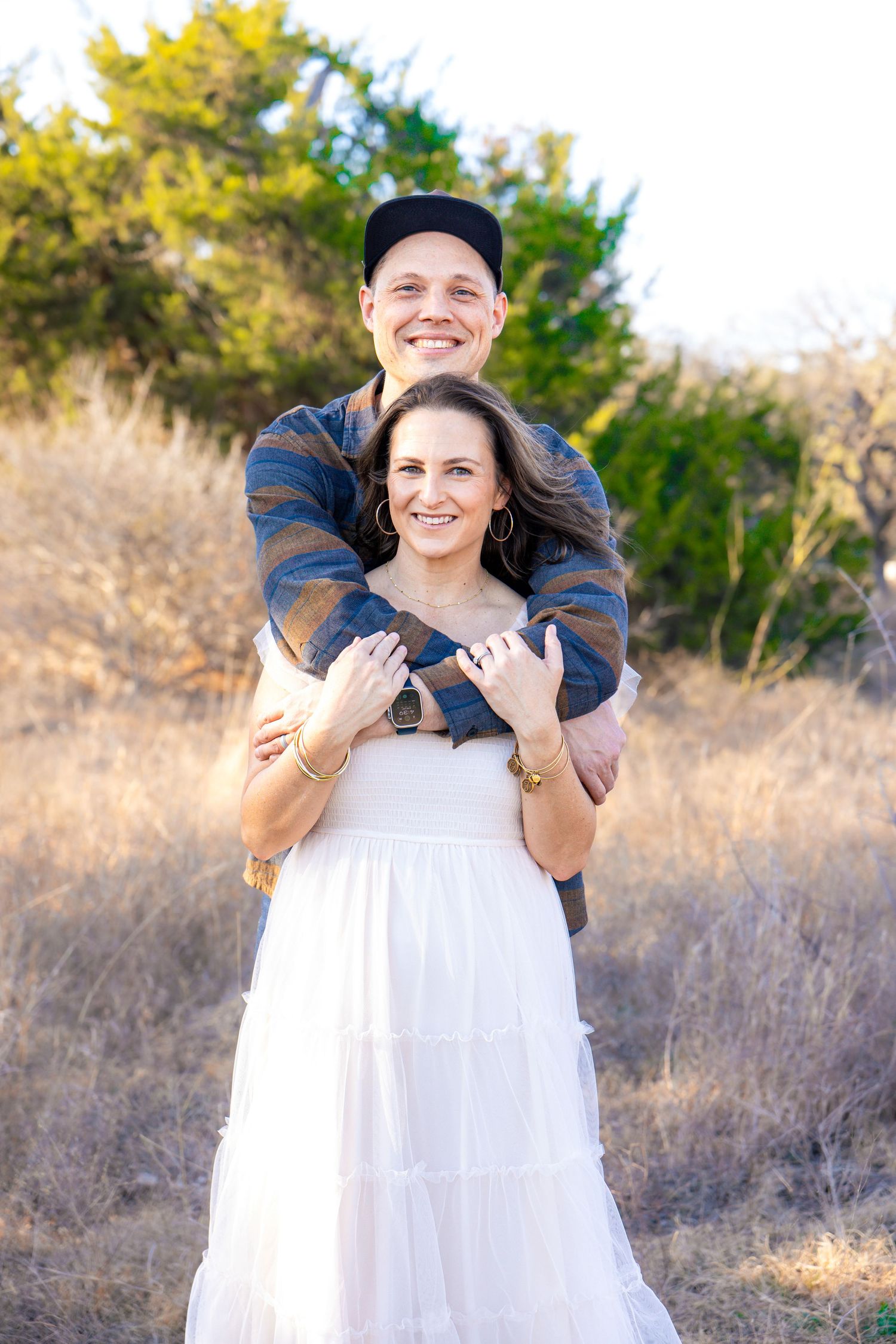 Happy couple embracing outdoors in a sunny field, woman in white dress with engagement ring, man in plaid shirt and cap.