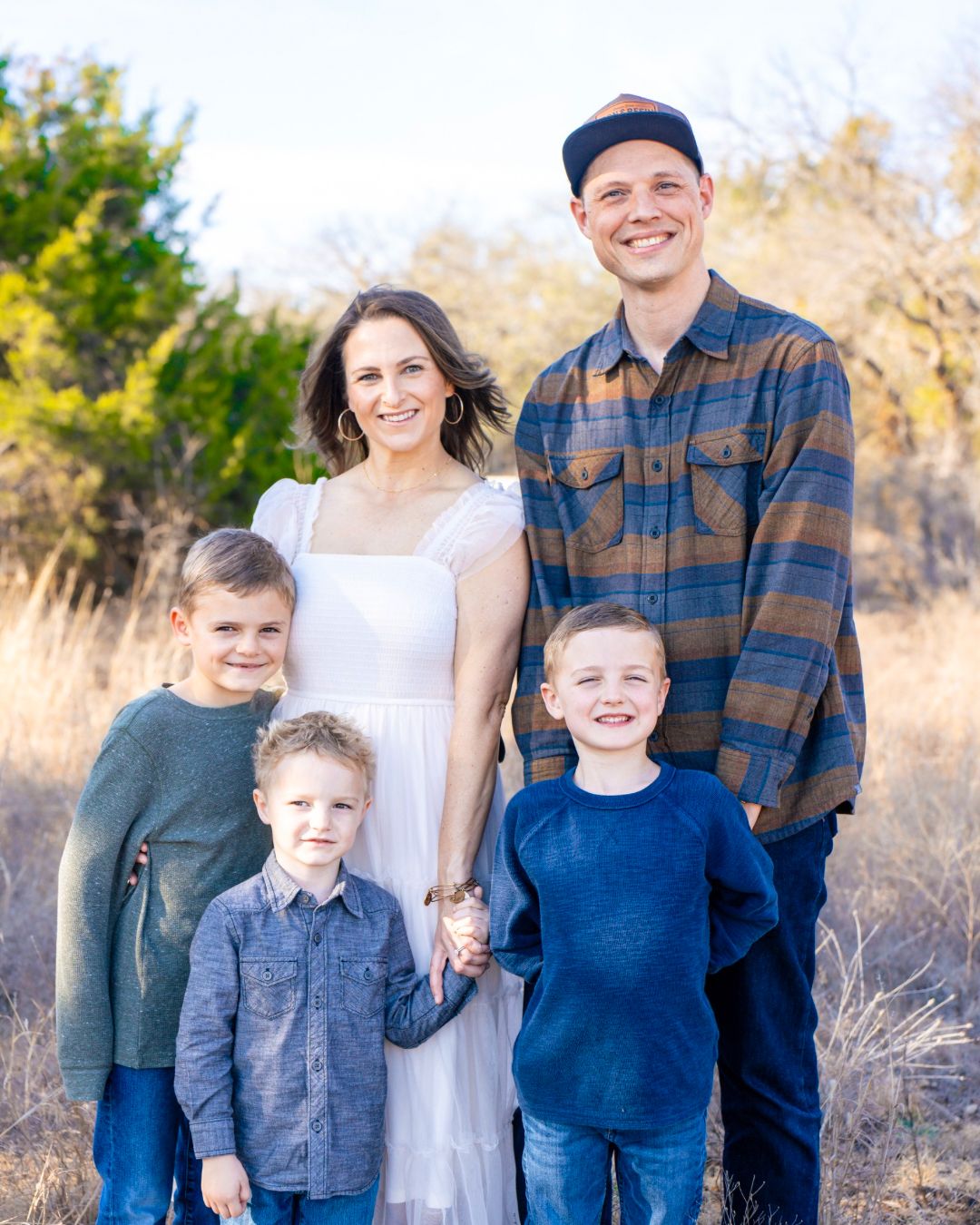 A happy family of five poses outdoors in a field, with parents and three young boys smiling in casual attire.