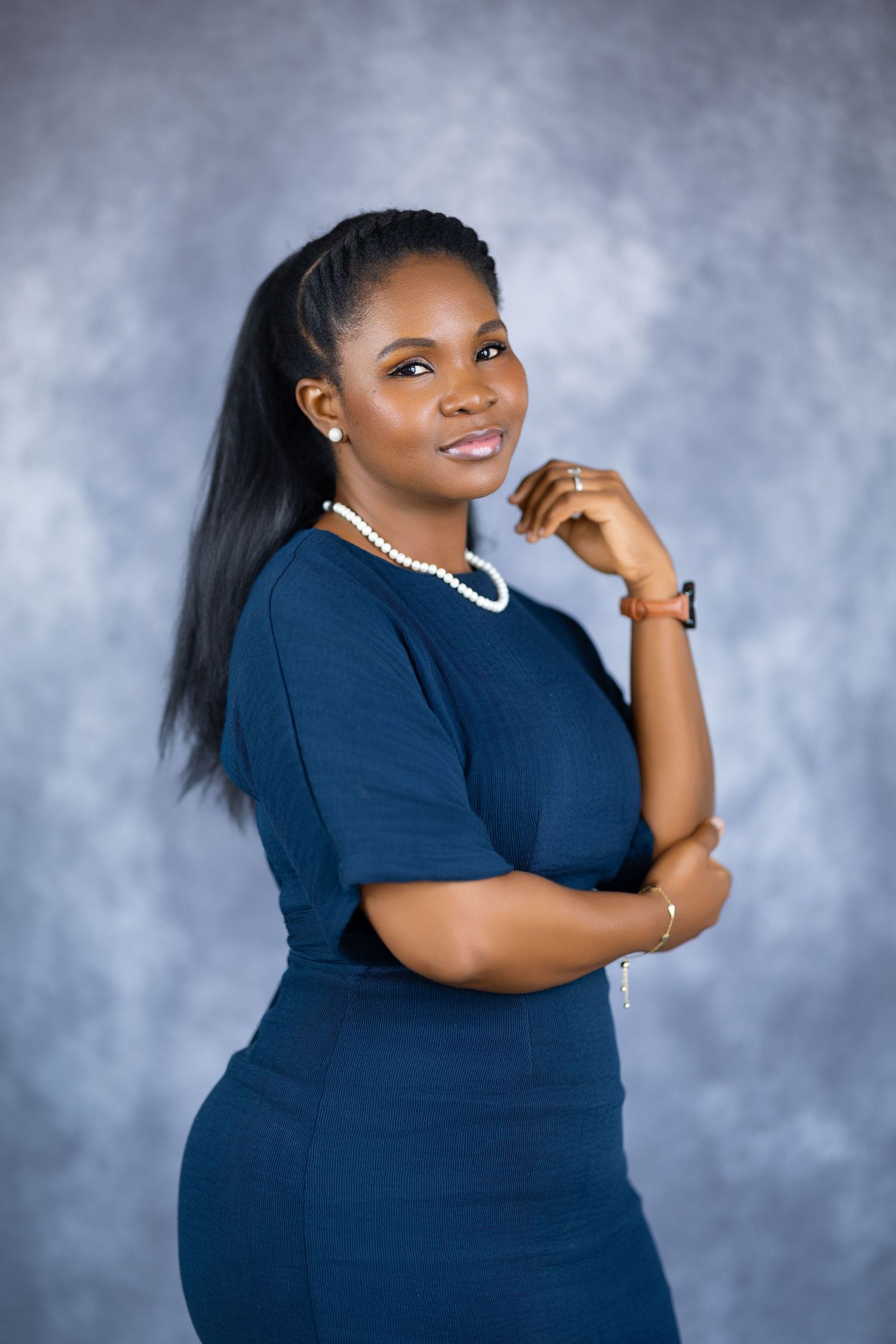 A young Black woman in a navy blue dress and pearl necklace poses confidently in a professional studio portrait.