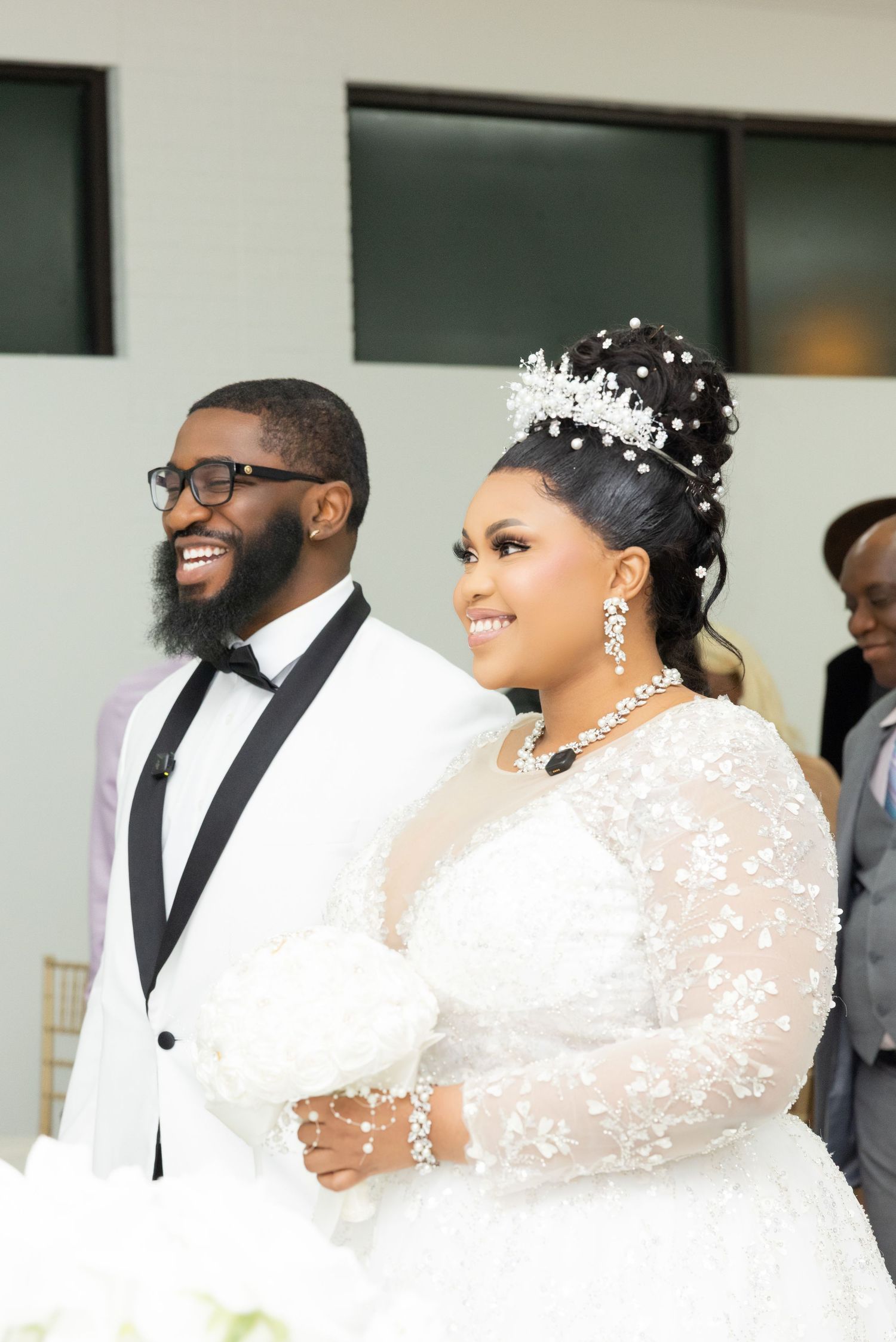 Happy couple at wedding ceremony, groom in white tuxedo, bride in lace gown with crystal crown.