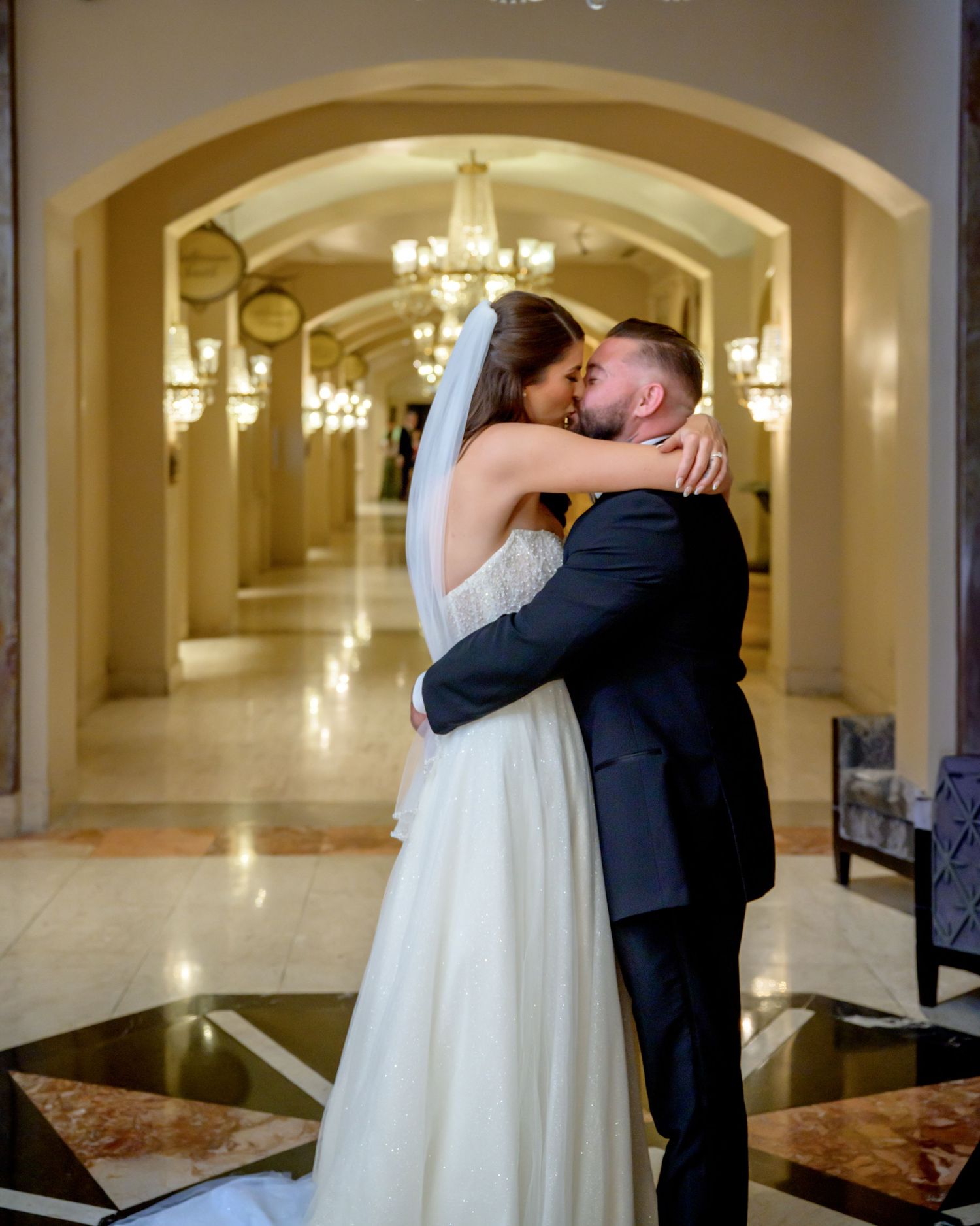A loving couple shares an intimate kiss in an elegant arched hallway with ornate chandeliers and marble floors.