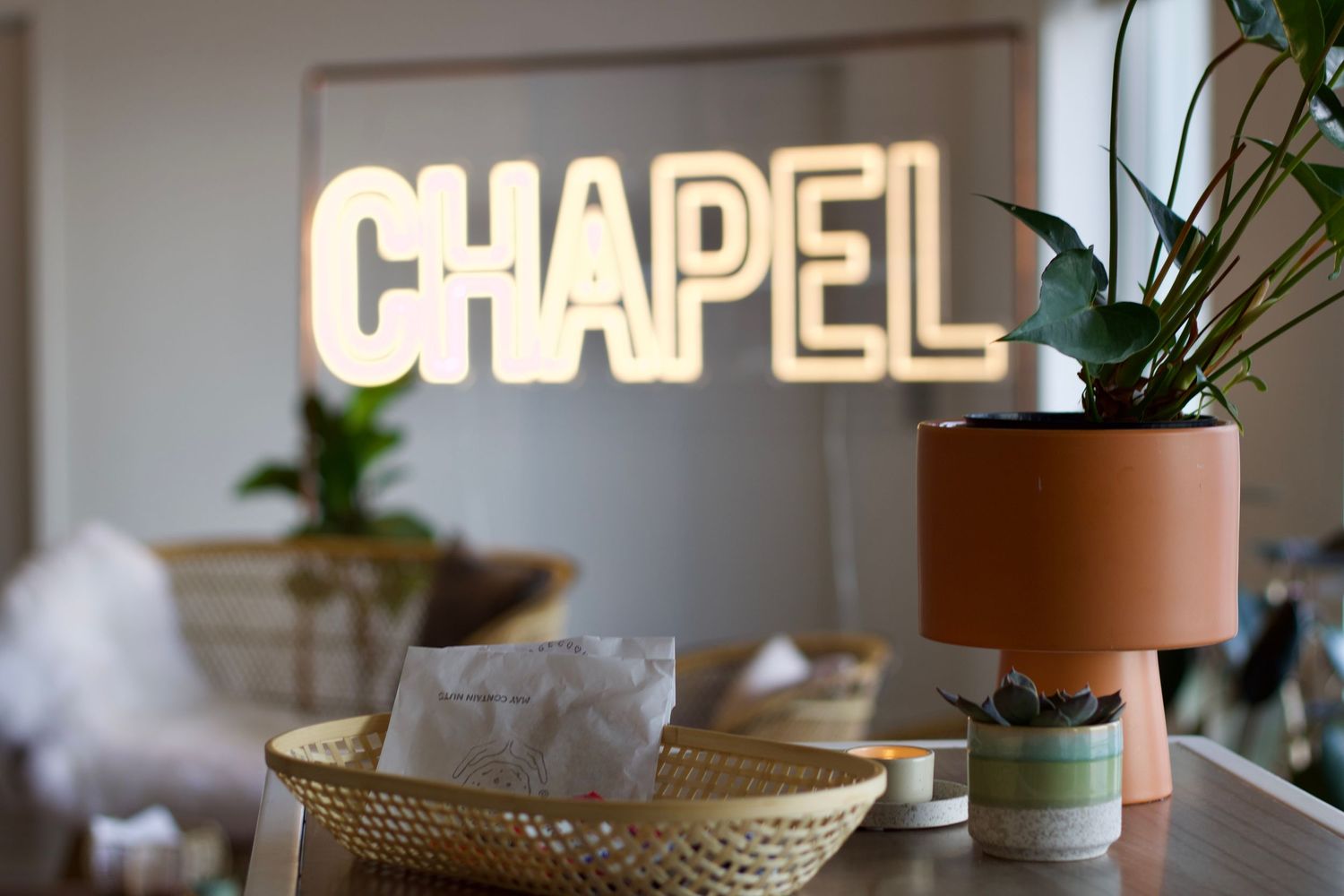 A glowing neon CHAPEL sign sits above a wicker basket and potted plant on a table.