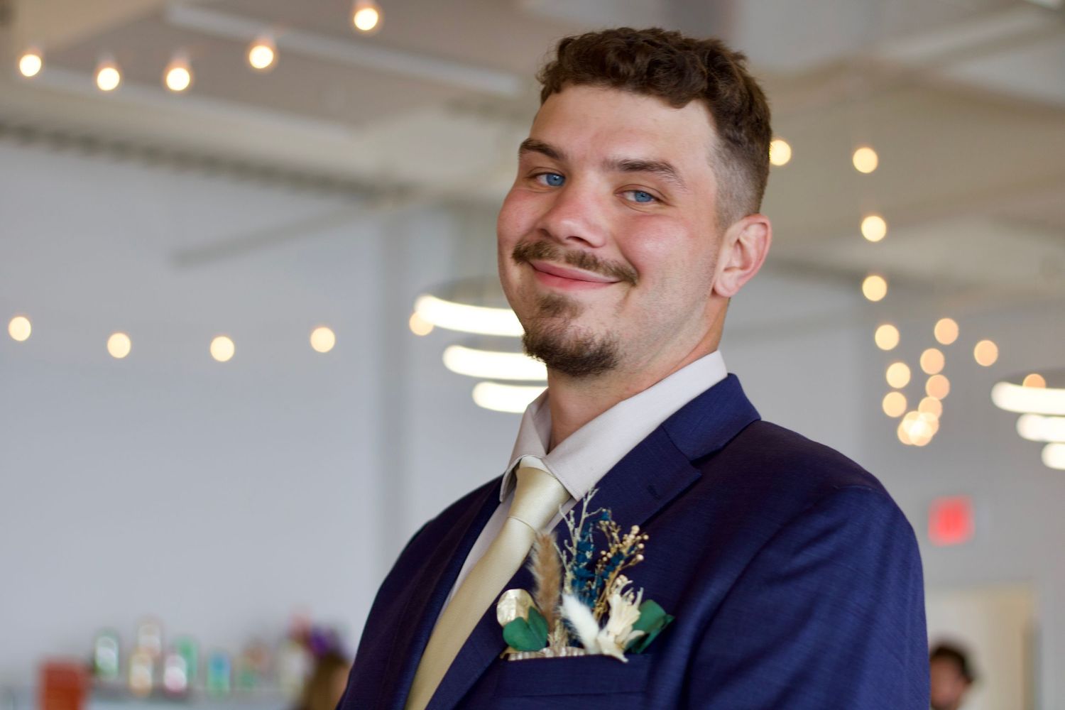 A young man in a navy suit with a boutonniere smiles at a wedding reception.