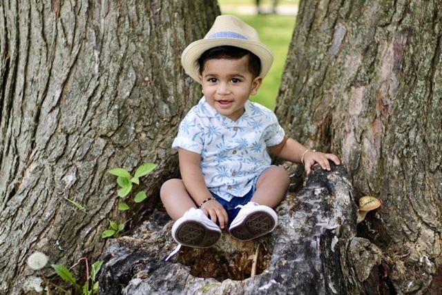 An adorable toddler boy in a straw hat sits smiling between tree trunks in a park.