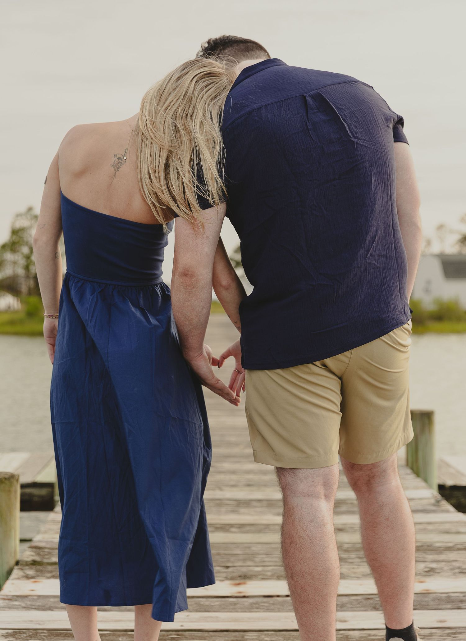Couple holding hands on a wooden dock overlooking calm water, woman in navy blue jumpsuit.