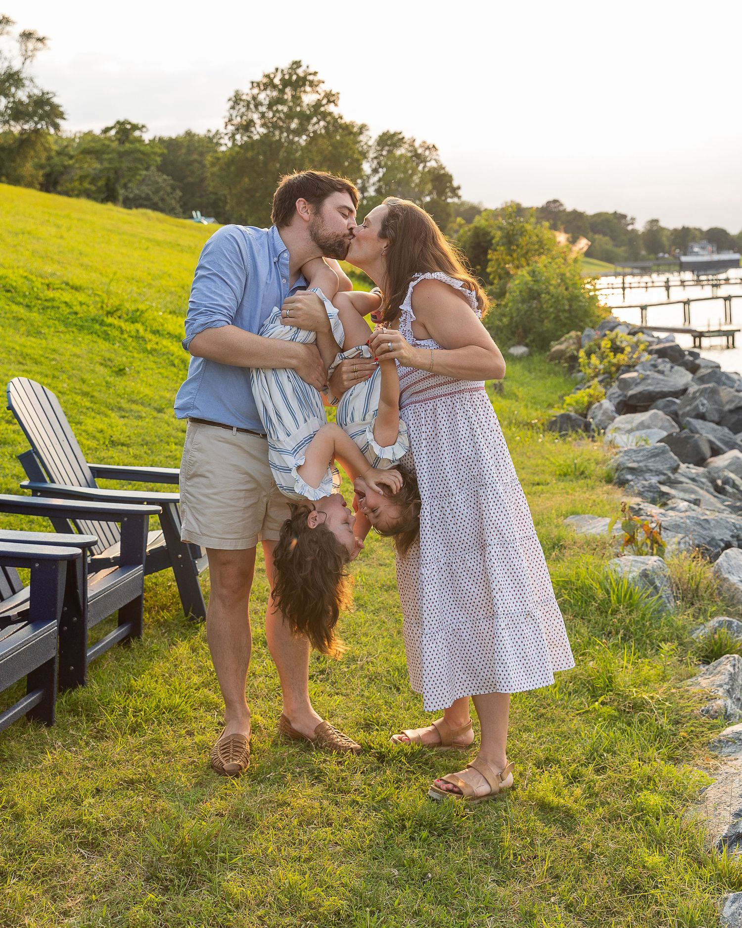 Family kissing outdoors by waterfront, parents holding child upside down on green lawn at golden hour.
