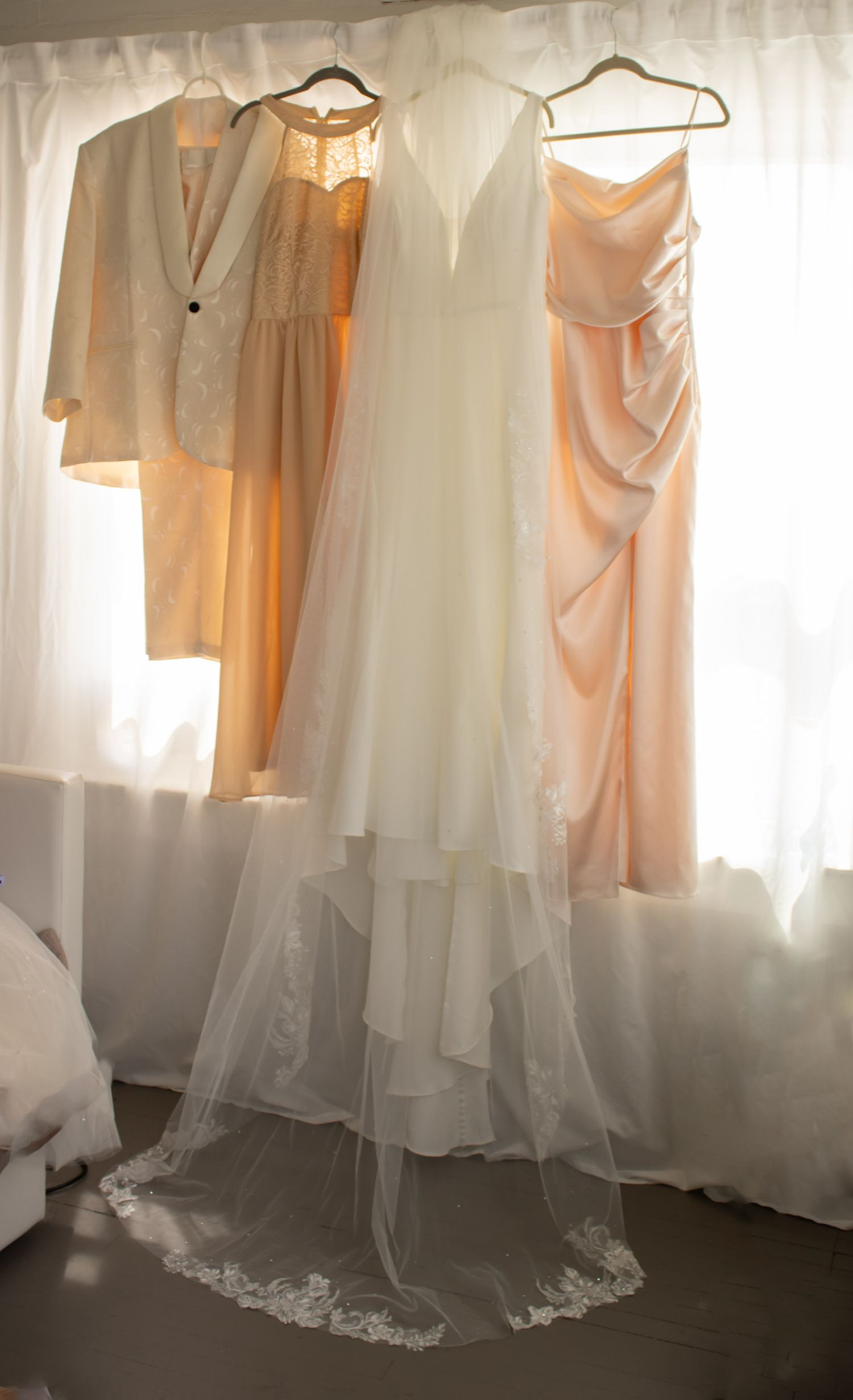 White wedding gown with lace-trimmed veil hanging beside peach bridesmaid dresses and a cream blazer on a window curtain rod.