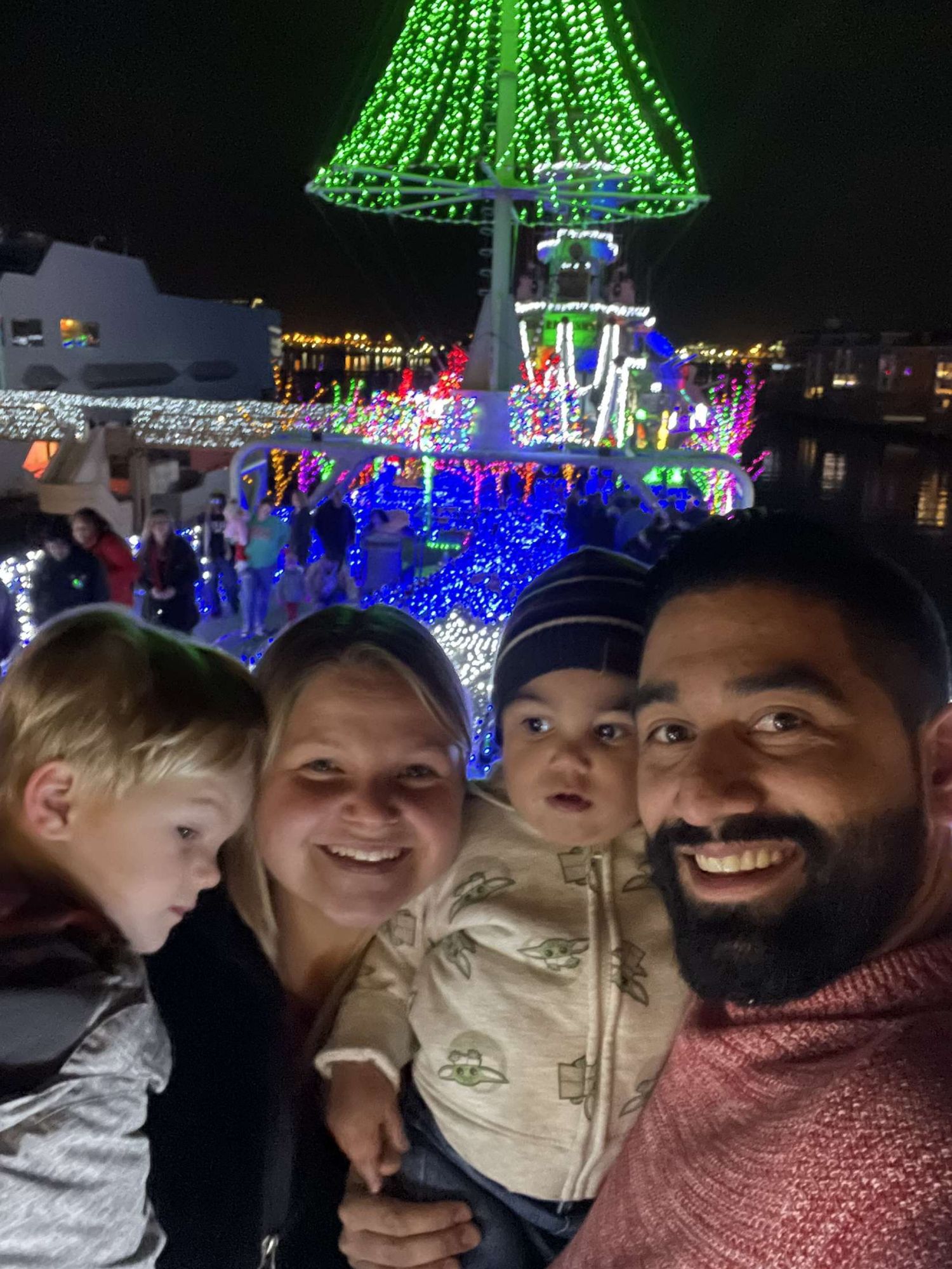 Family of four smiling for selfie at colorful Christmas light display at night.