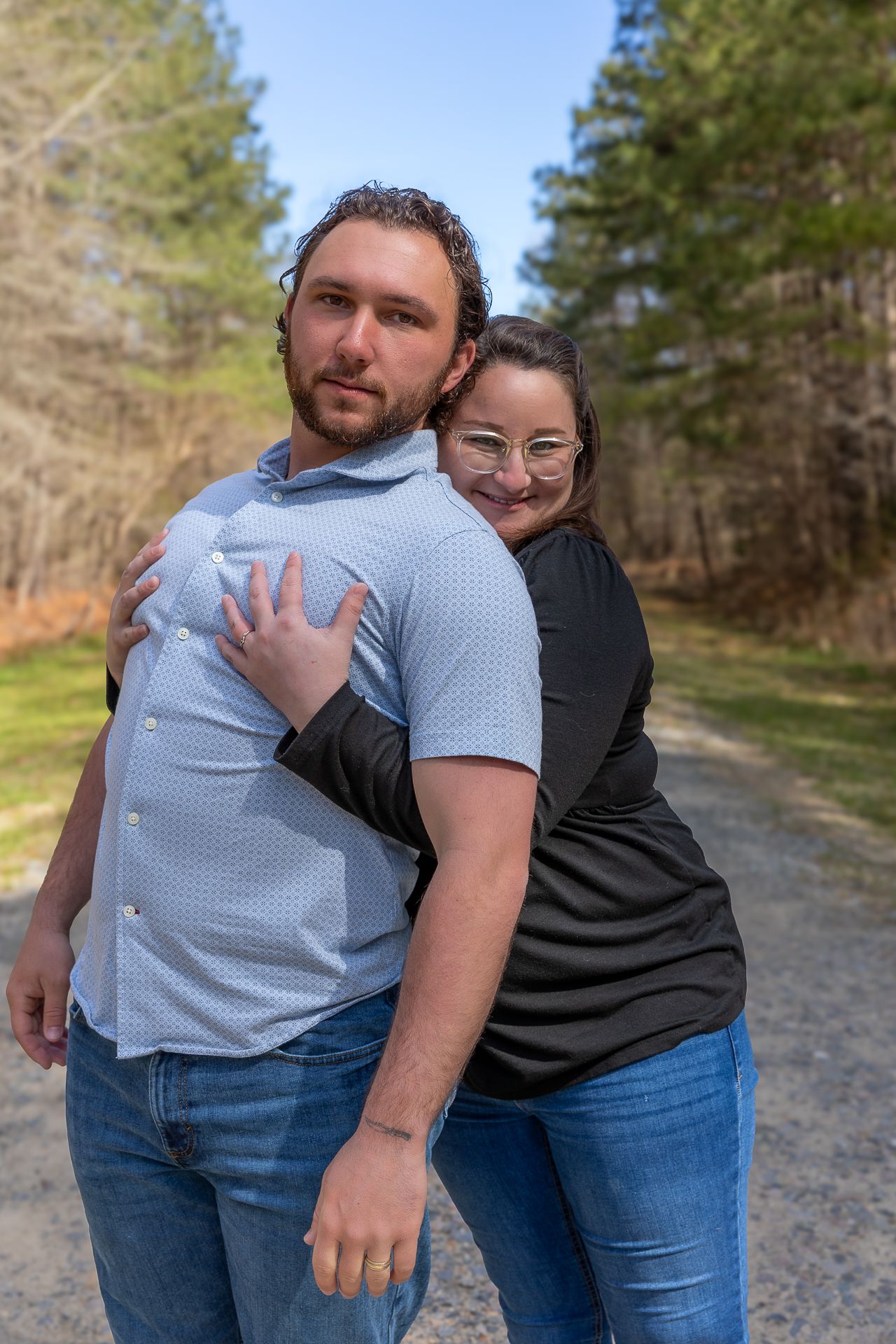 Couple posing on a wooded gravel path, woman hugging man from behind, both smiling, wearing casual clothes outdoors.