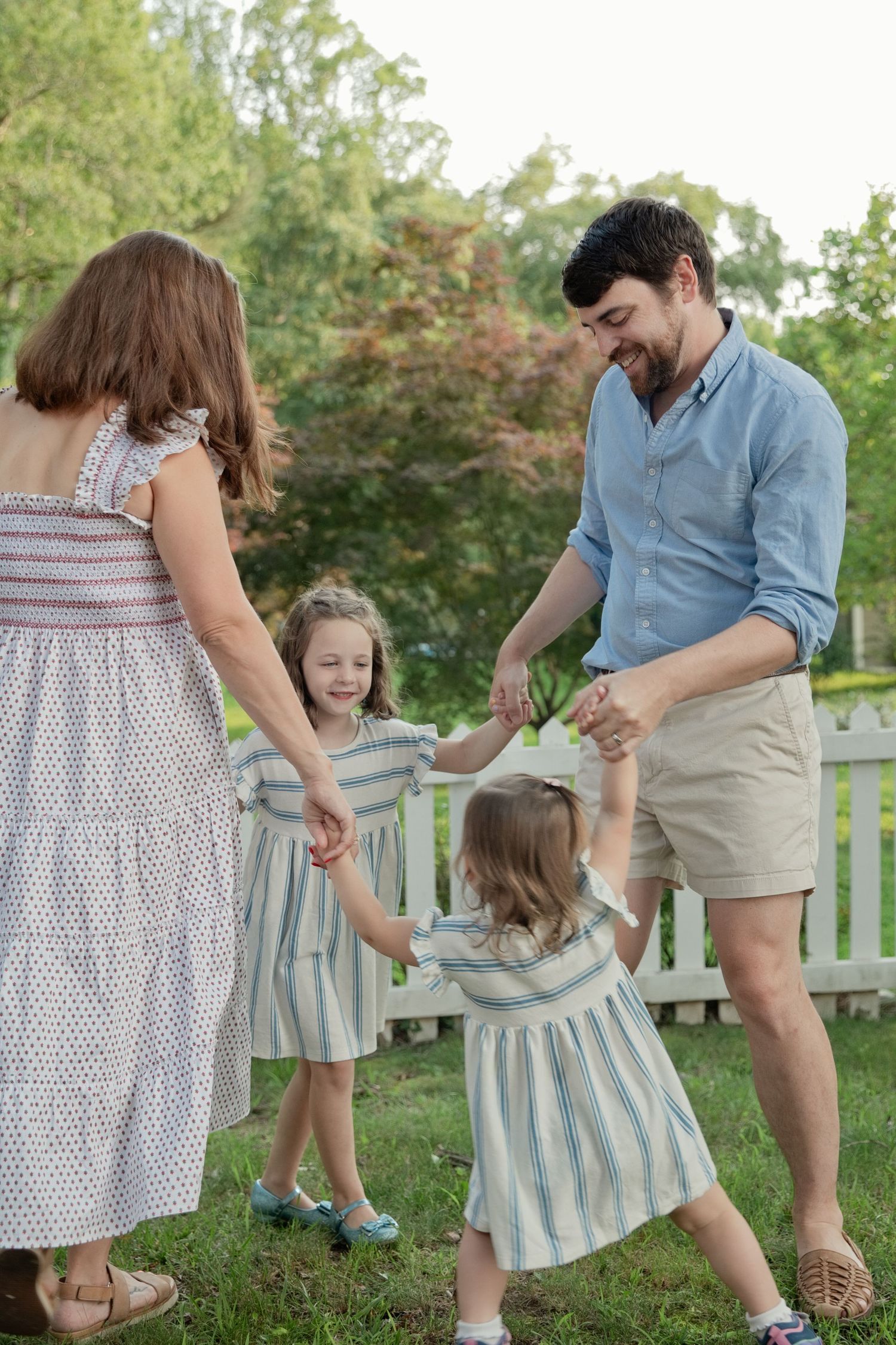 A joyful family of four holds hands and dances together in a lush green backyard on a sunny day.