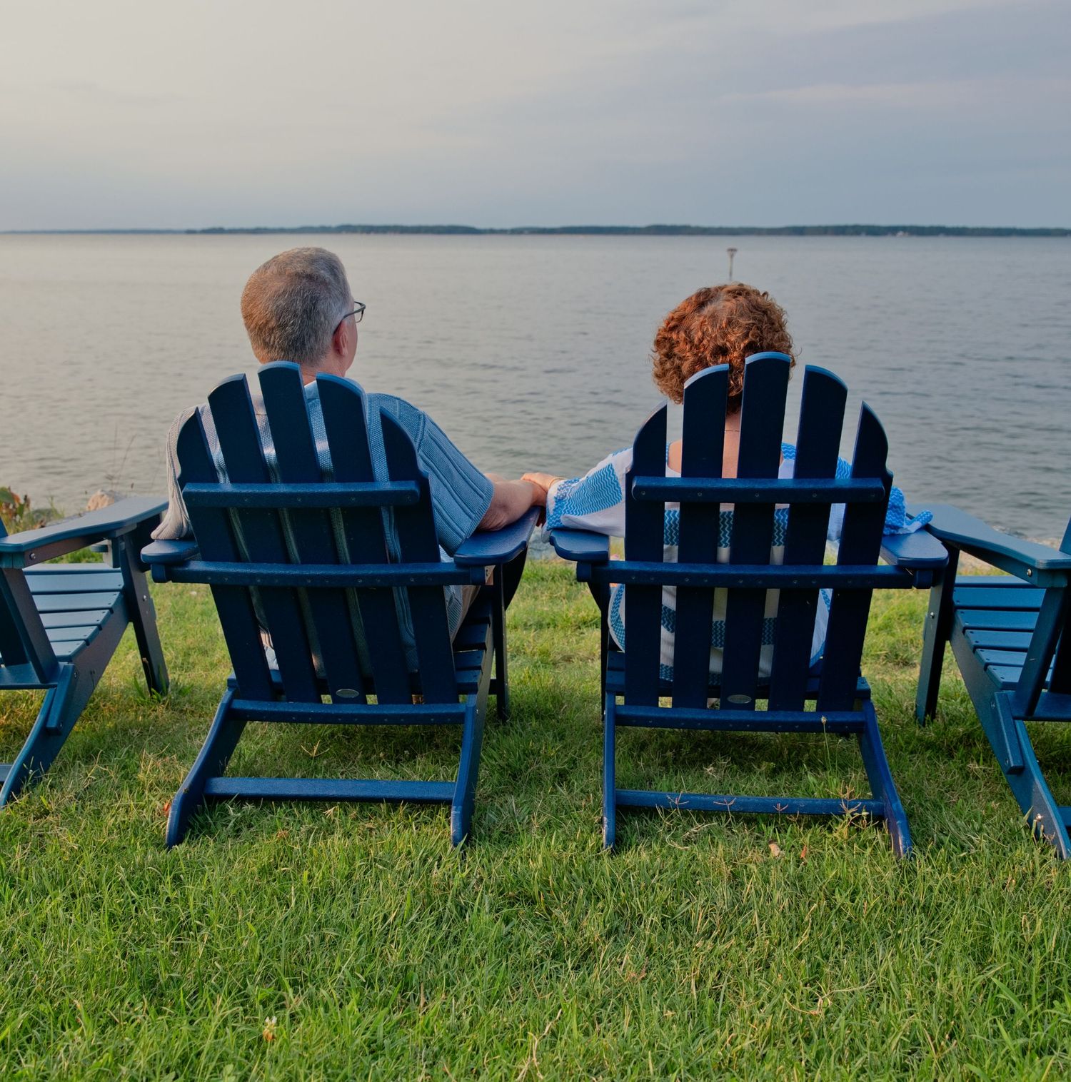 A couple relaxes in blue Adirondack chairs overlooking a serene lake at dusk on a grassy shore.