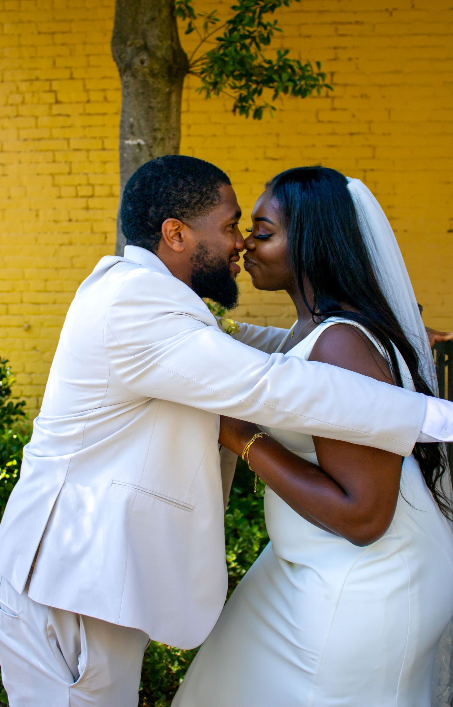 A bride and groom share a romantic kiss outdoors, both dressed in white, against a vibrant yellow brick wall.