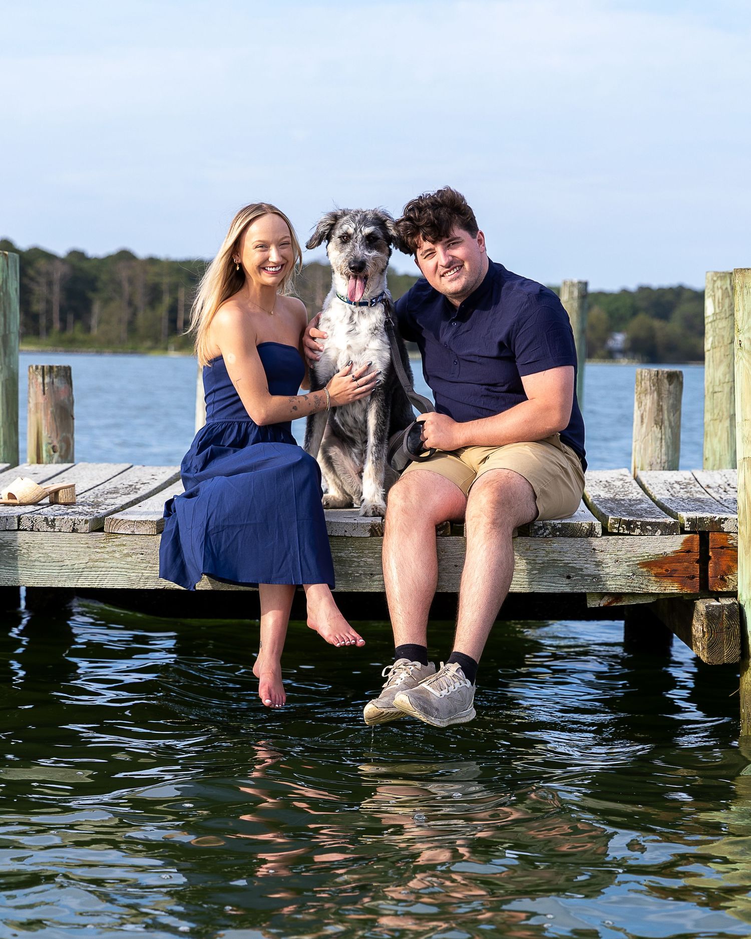 Couple and their dog sitting on a wooden dock over water, woman in navy dress, man in navy polo and khaki shorts.