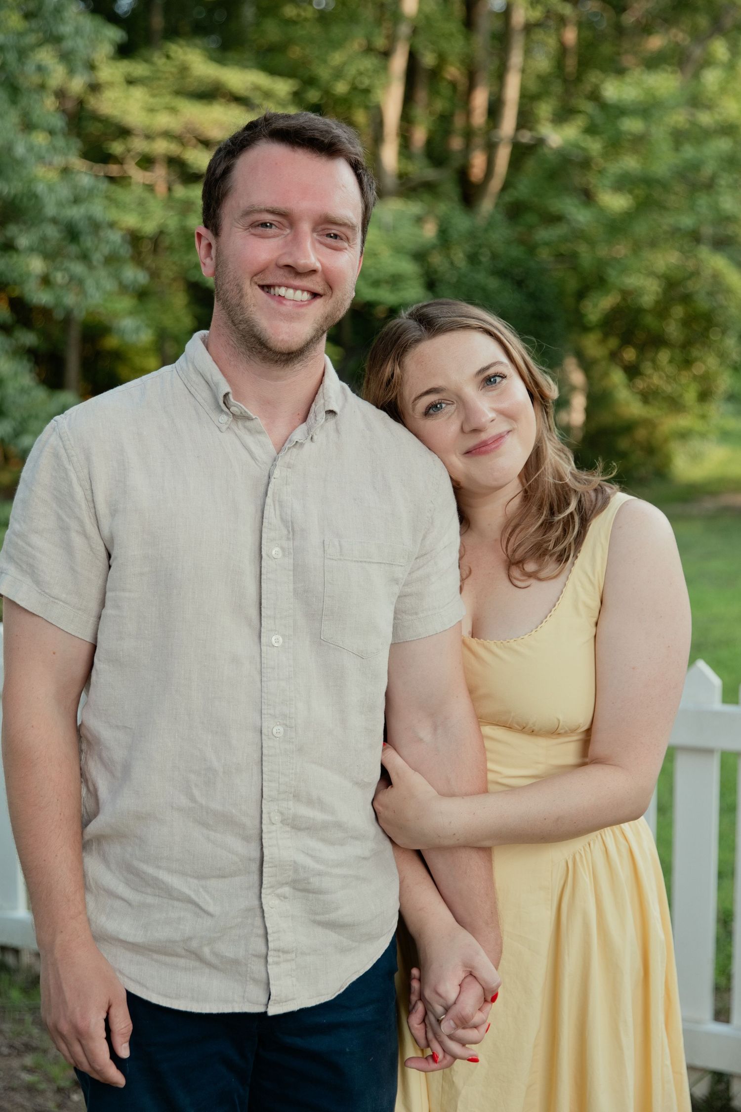 Smiling couple posing together outdoors, woman in yellow dress and man in beige linen shirt, with green trees in background.
