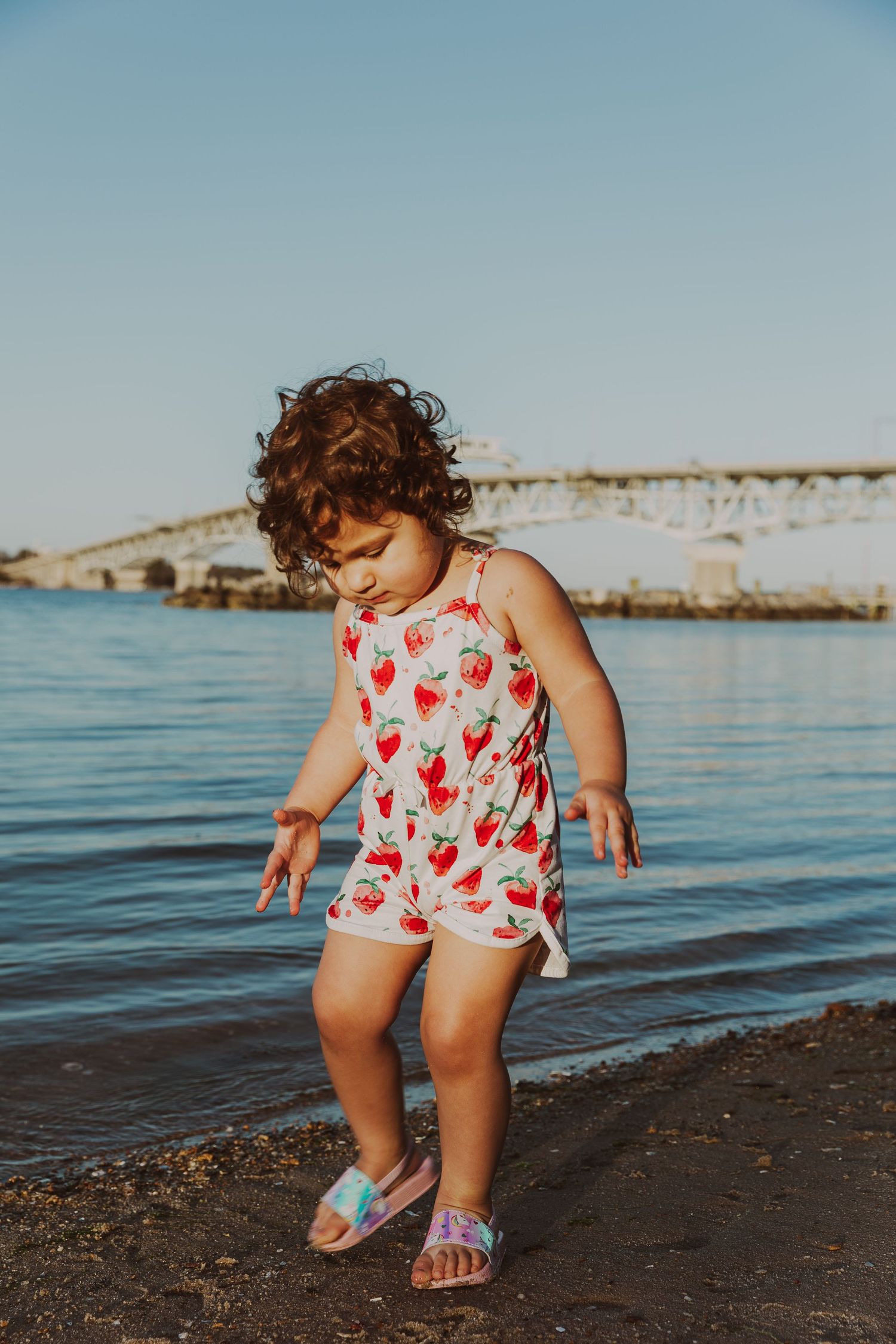 A toddler in a strawberry romper plays near the water's edge with a bridge in the background.