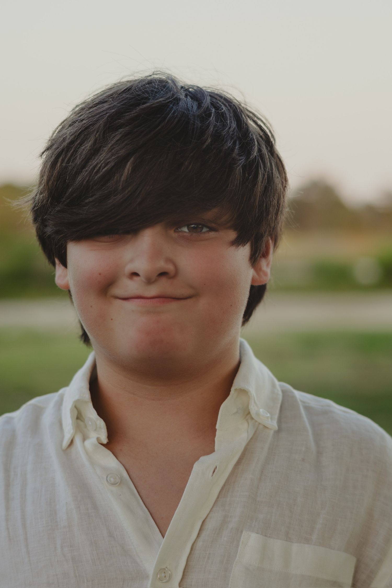 A young boy with dark hair smiles softly, wearing a white linen shirt outdoors in a green field.