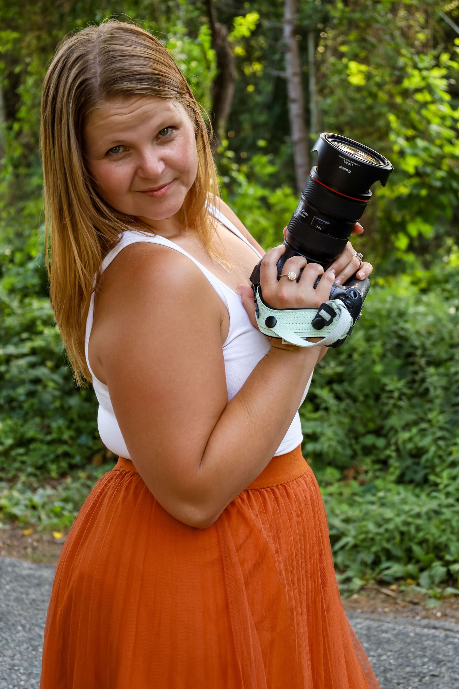 A smiling female photographer in an orange skirt holds a professional DSLR camera outdoors.