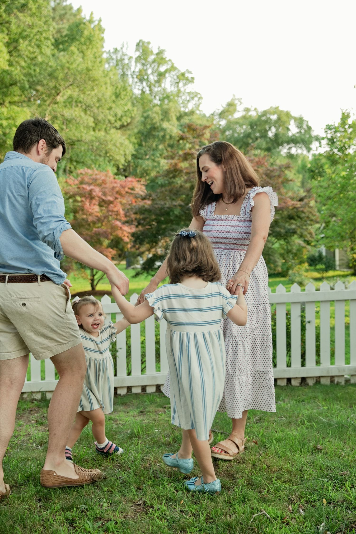 A joyful family of four holds hands and dances together in a lush green backyard with a white picket fence.