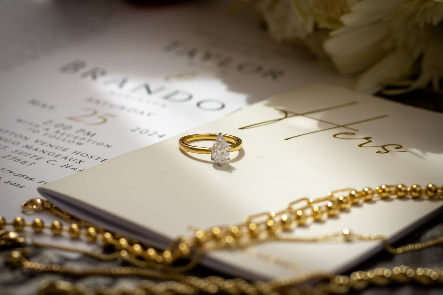 Gold diamond engagement ring resting on elegant wedding invitation with pearl necklace and white flowers in background.