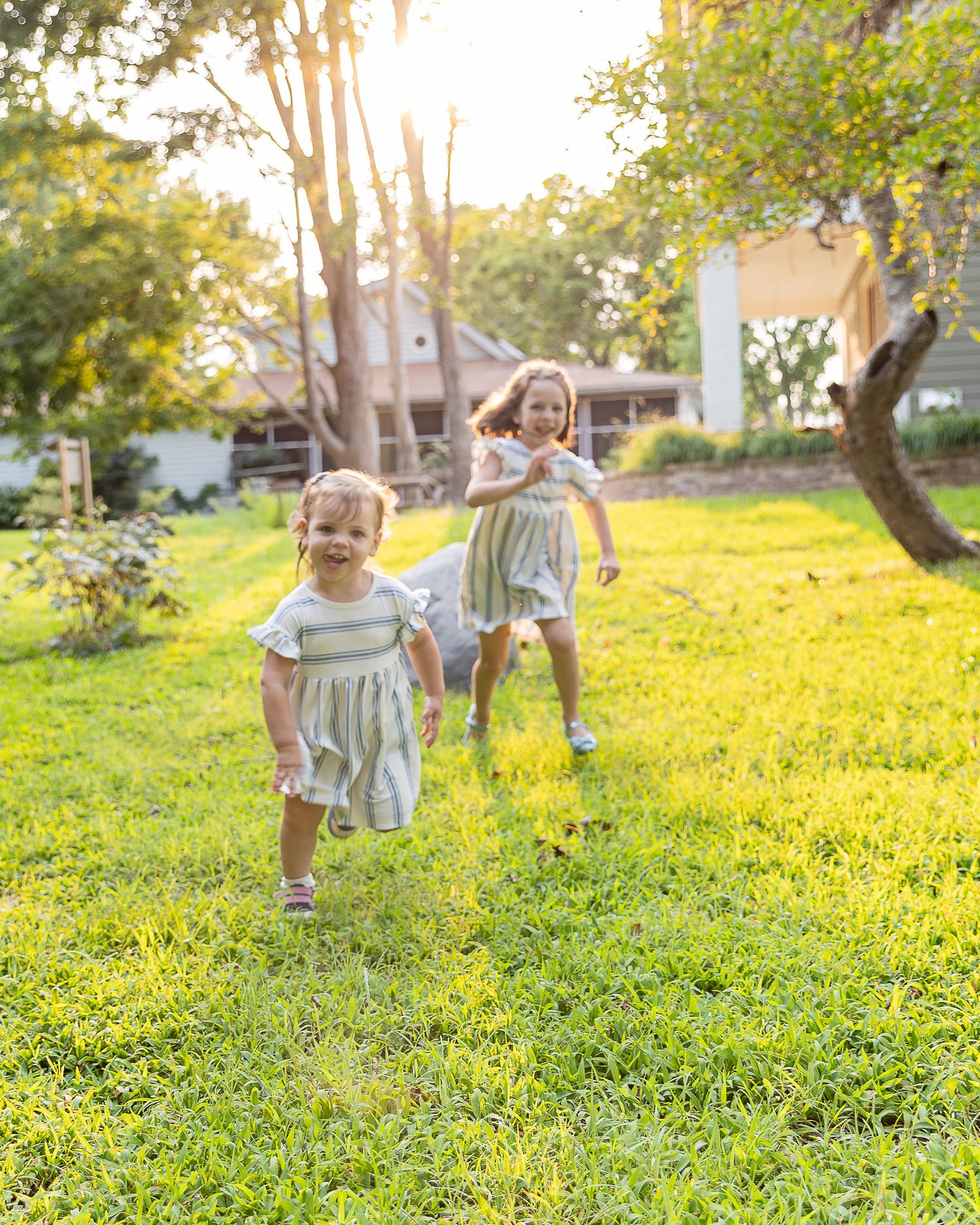 Two young girls in matching dresses running and playing on lush green grass in a sunny backyard.