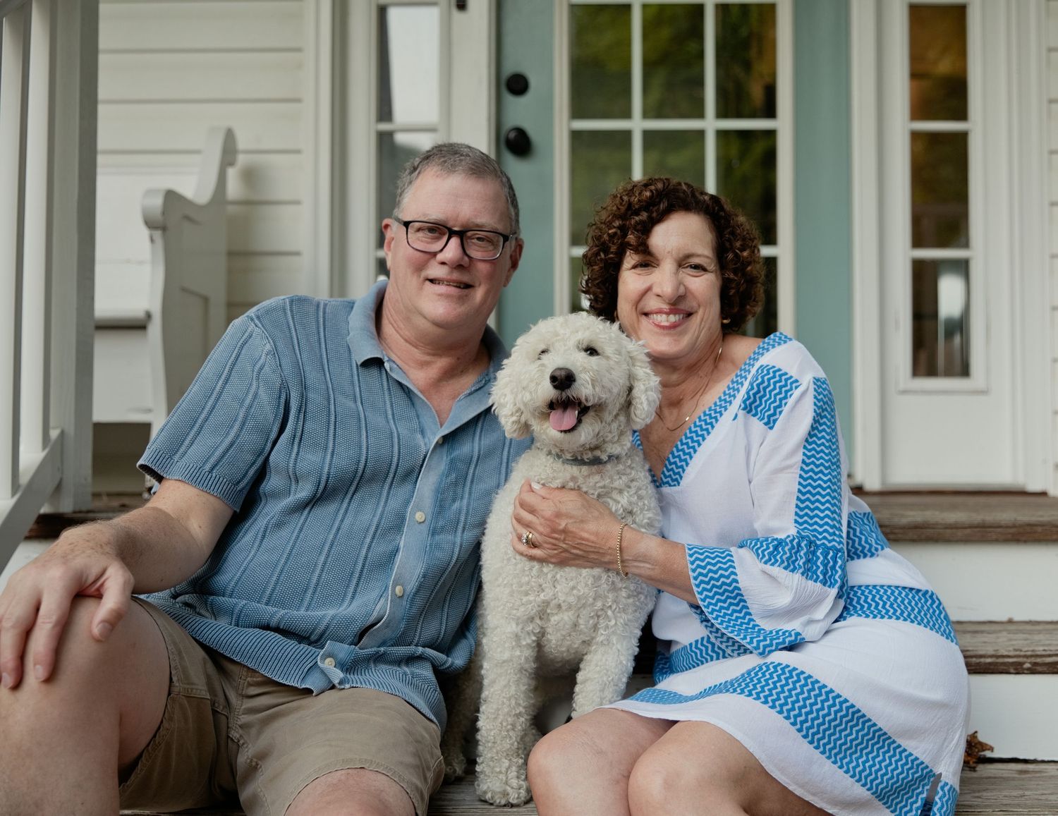 A smiling couple sits on porch steps holding a fluffy white dog outside a charming home.
