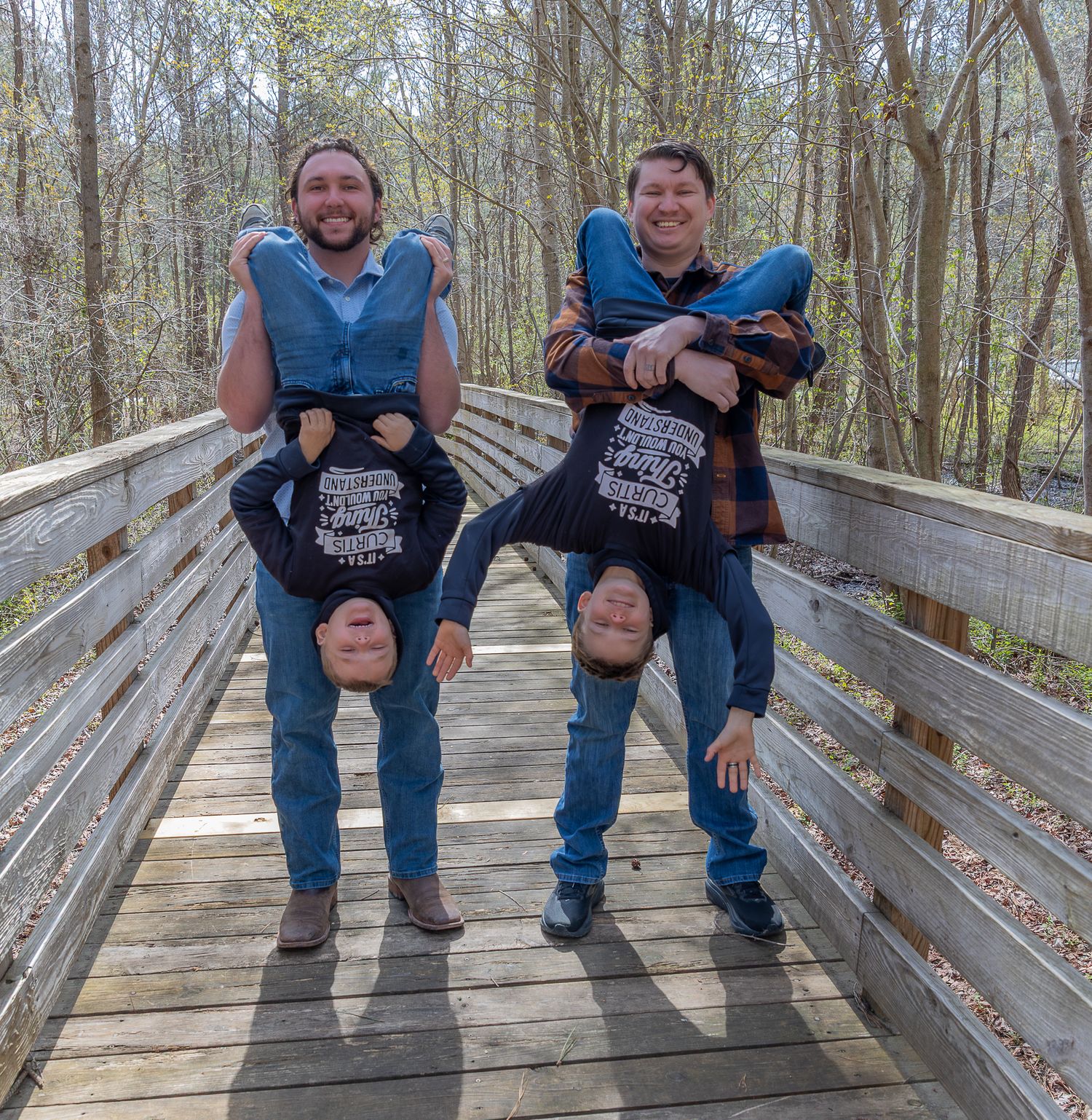 Two men carry people upside down on a wooden bridge surrounded by autumn trees, smiling for a fun outdoor photo.