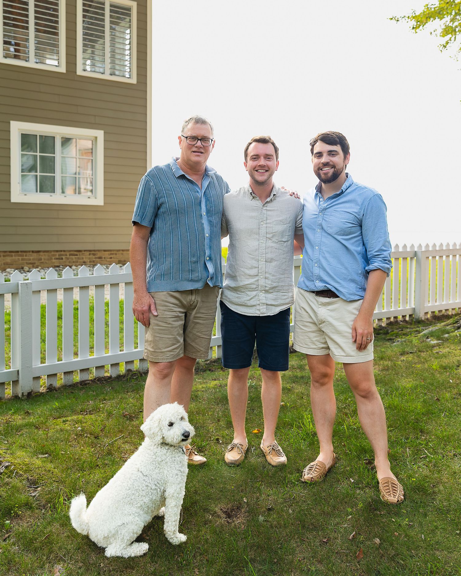 Three smiling men stand together in a backyard with a white fluffy dog beside them.