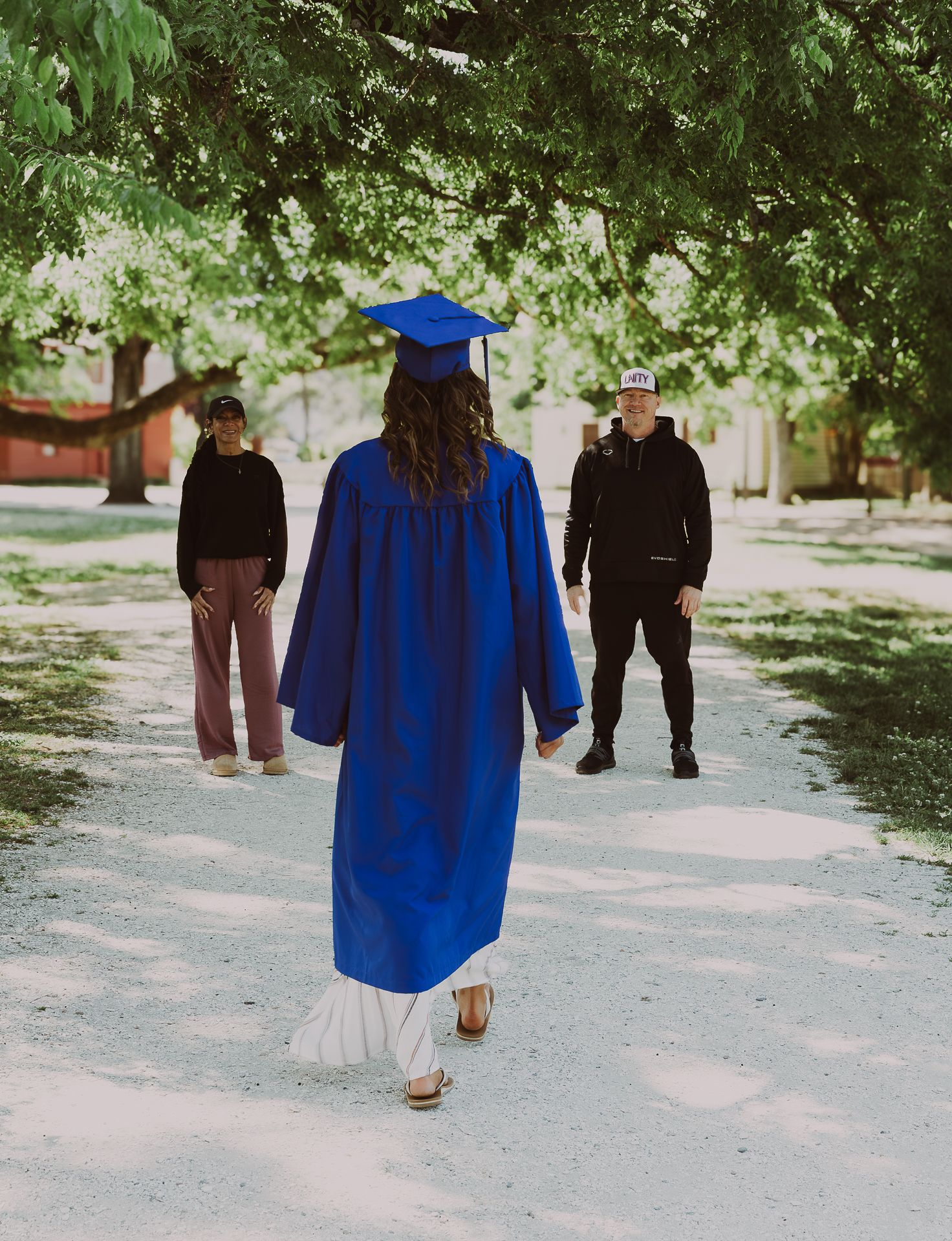 Graduate in blue cap and gown walks toward two people on a tree-lined path during outdoor graduation celebration.