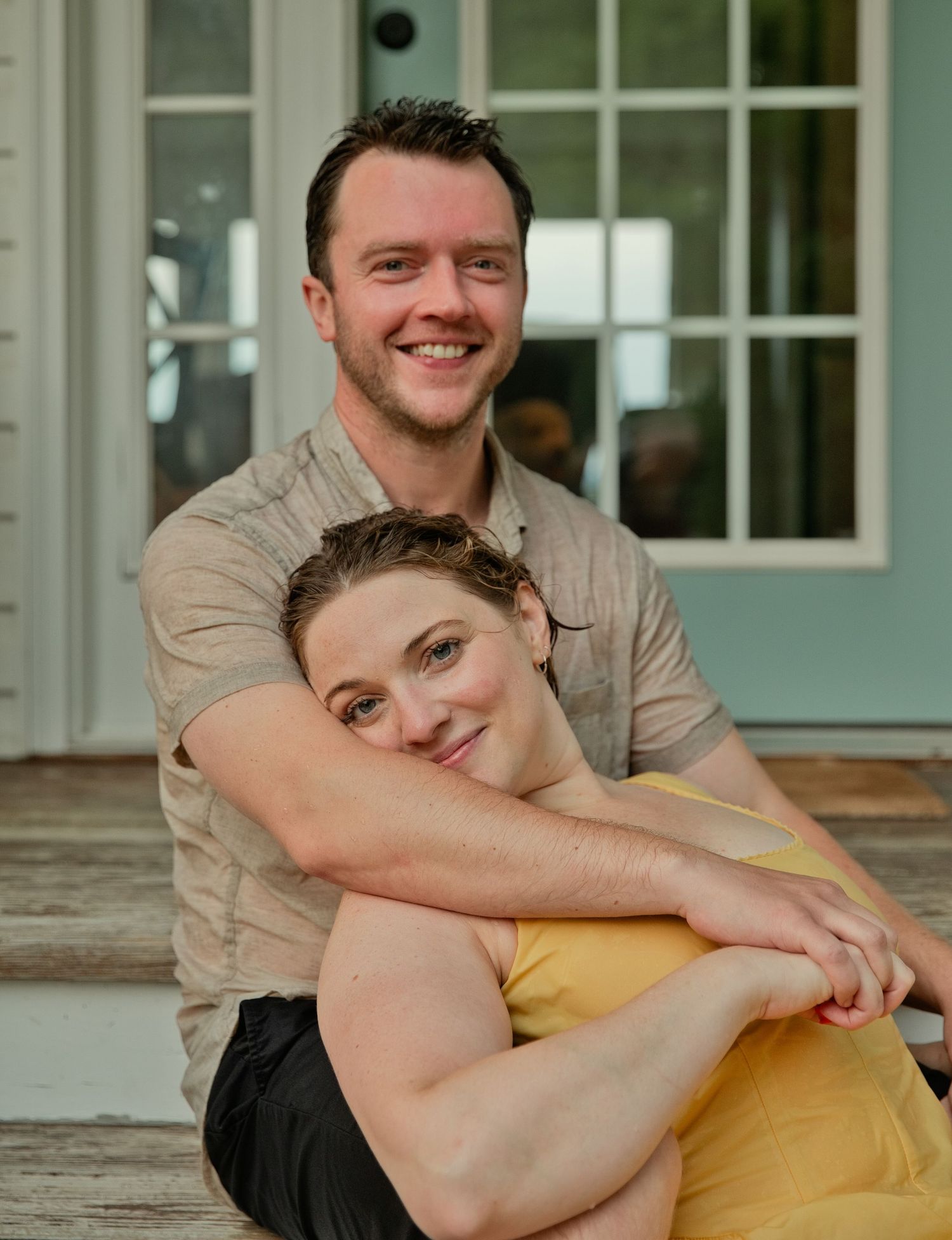 A smiling couple poses warmly together on a porch, woman in yellow leaning against mans chest.