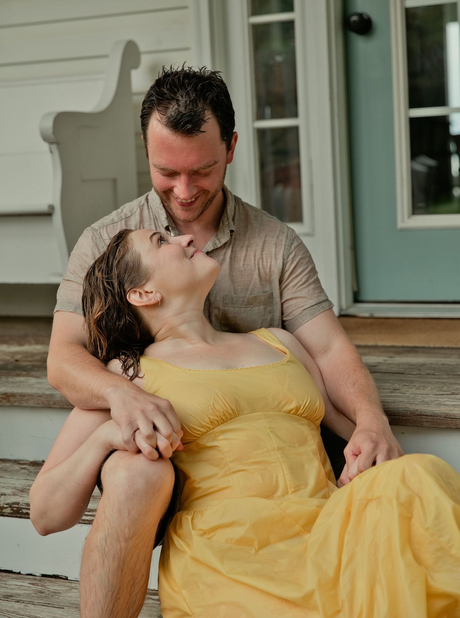 A smiling couple shares a tender moment on wooden porch steps, woman in yellow dress leaning back.
