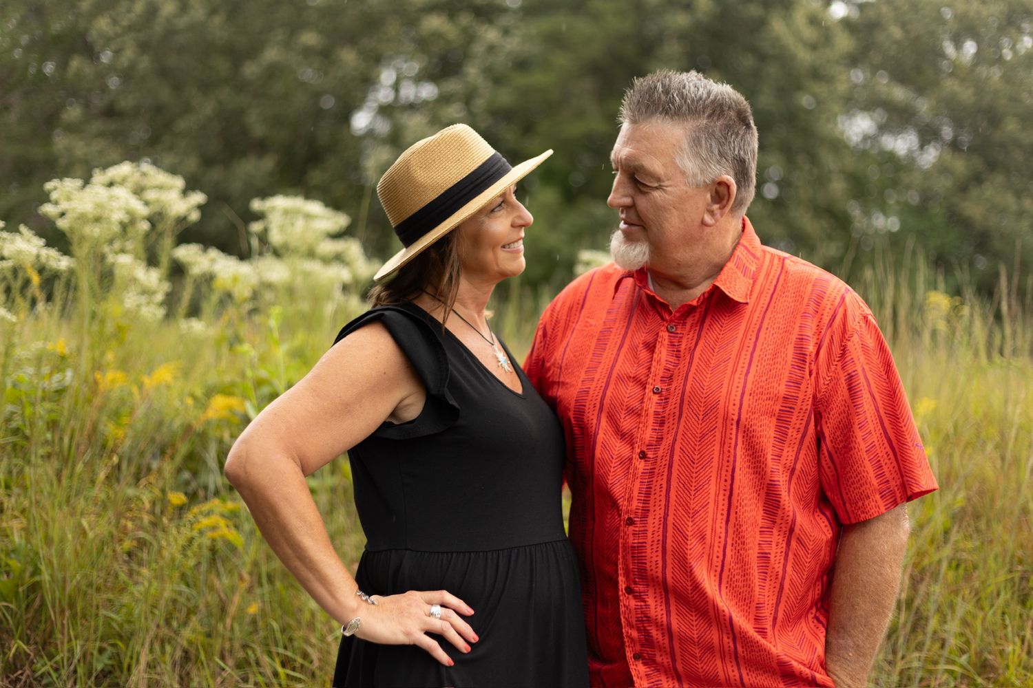 A smiling middle-aged couple stands together in a wildflower meadow, the woman wearing a straw hat and black dress.