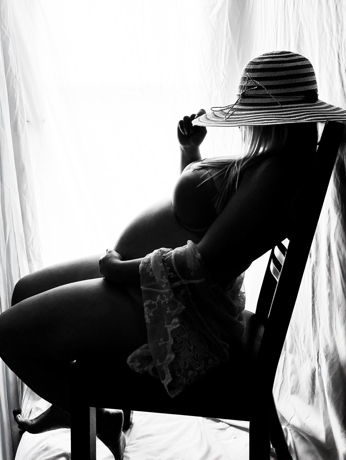 Black and white maternity photo of a pregnant woman in a floppy striped hat sitting gracefully on a chair by sheer curtains.