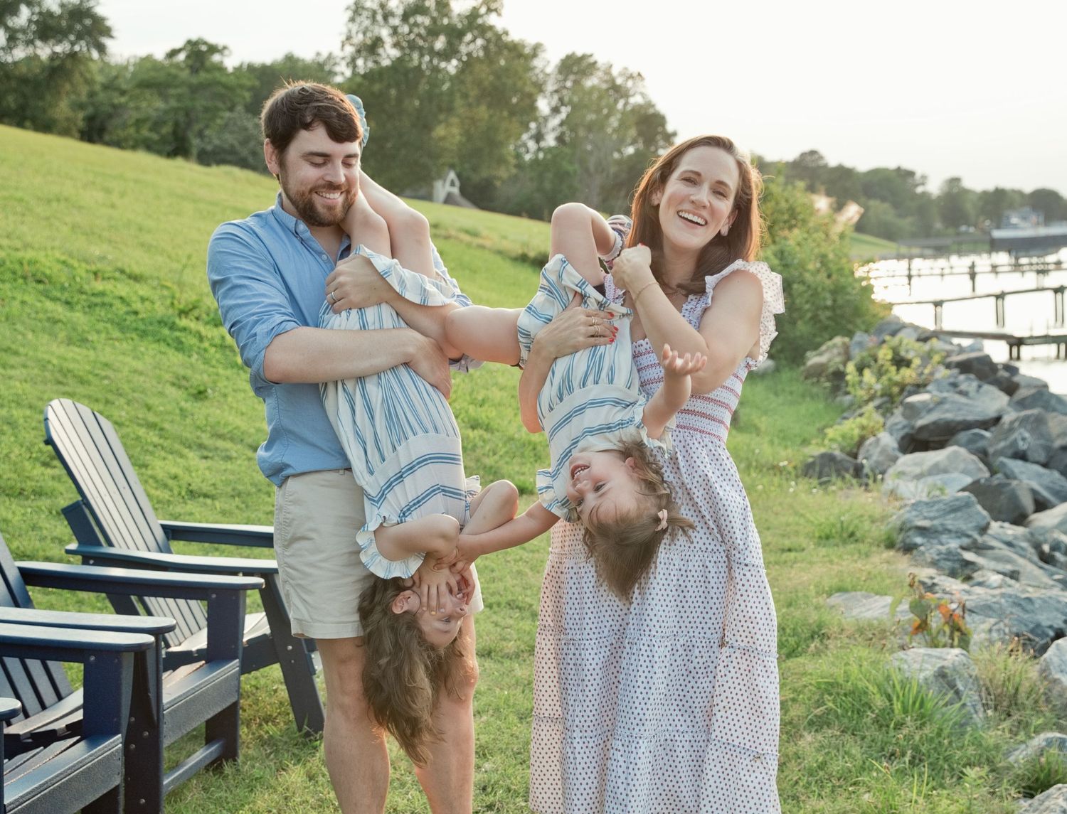 A laughing family of four playfully holds two young girls upside down near a scenic waterfront.