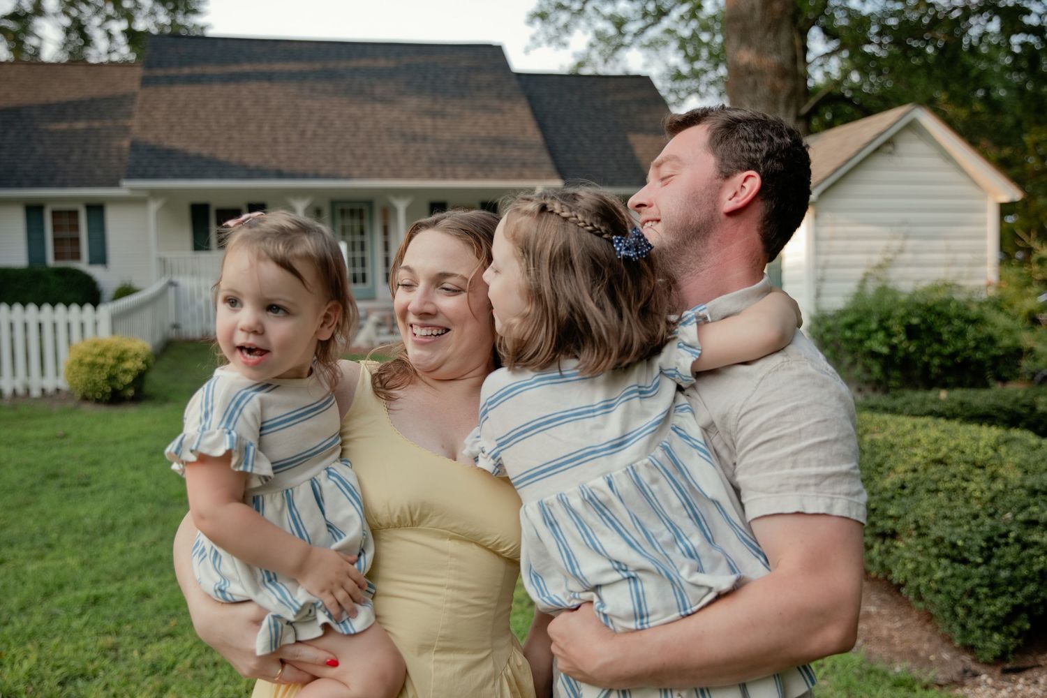 Happy family with two young girls in matching outfits hugging parents in front of a white house.