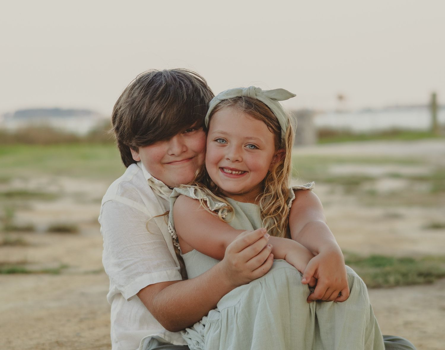 Two young siblings share a warm hug outdoors, smiling joyfully in soft natural light.