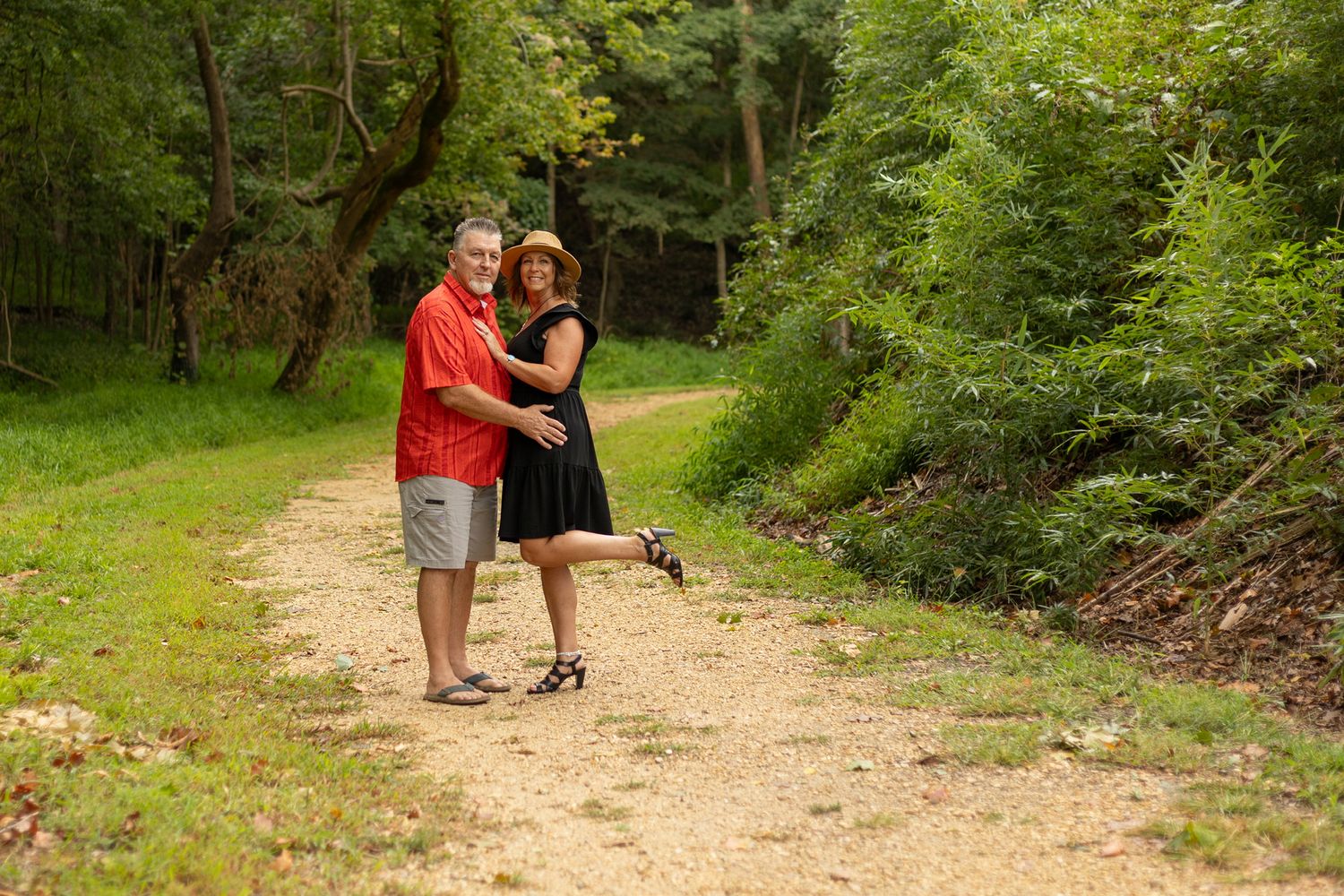 Couple posing on a scenic forest trail surrounded by lush green trees on a sunny day.