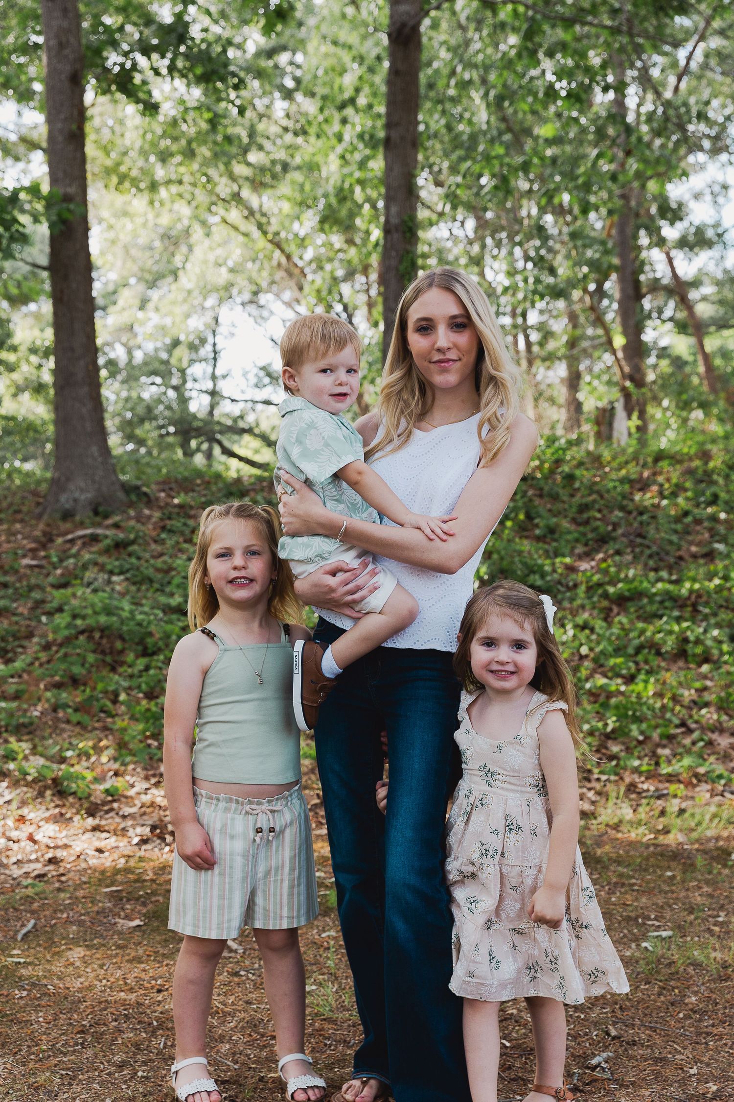 A mother holds her toddler son while posing with two young daughters outdoors in a wooded park setting.