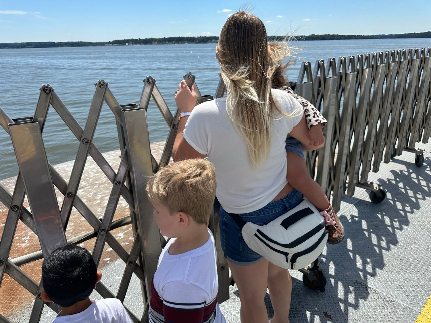 A woman and two children stand at a waterfront fence enjoying the sunny ocean view.