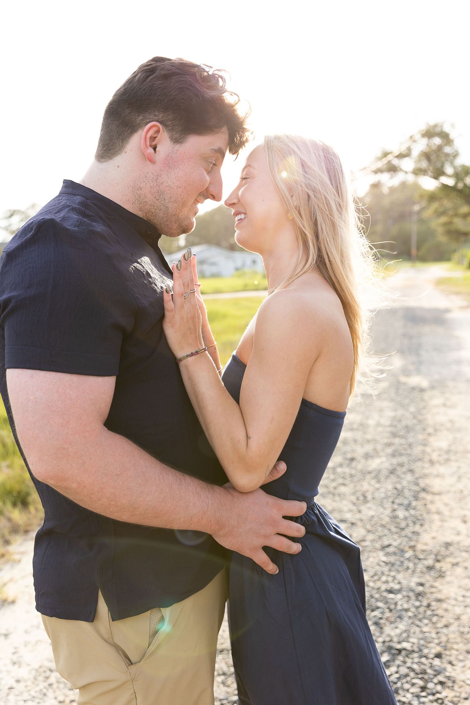 A couple shares a romantic moment on a sunlit gravel road, smiling at each other in coordinating navy outfits.