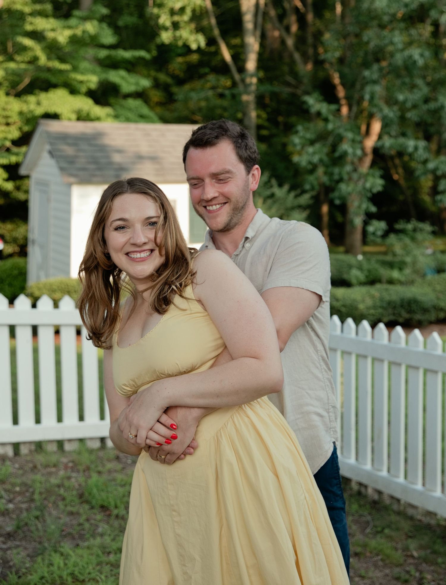A smiling couple embraces in a lush backyard with a white picket fence, woman in yellow dress.
