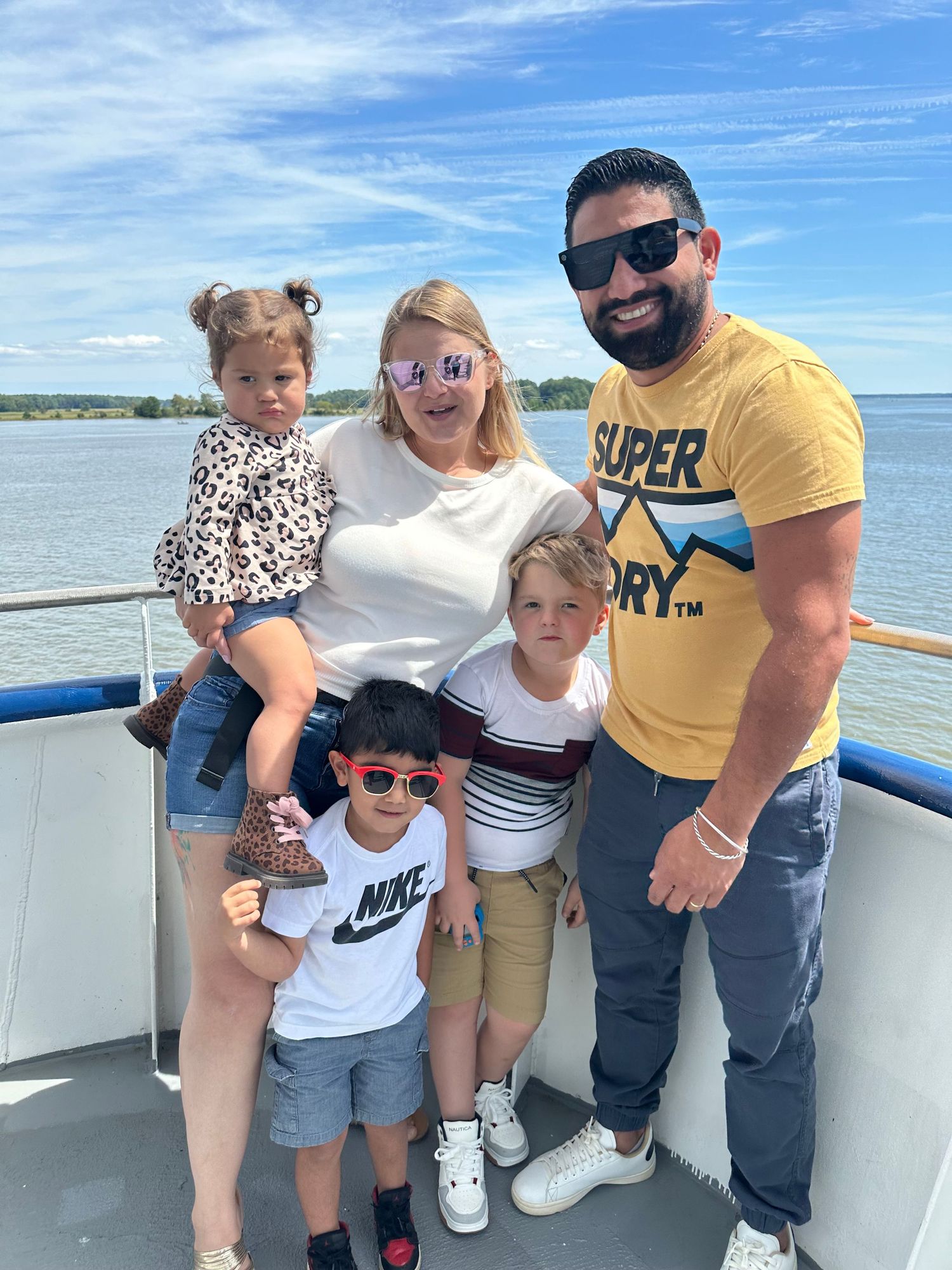 Family of five posing on a boat deck on a sunny day, with calm blue waters and green shoreline in the background.