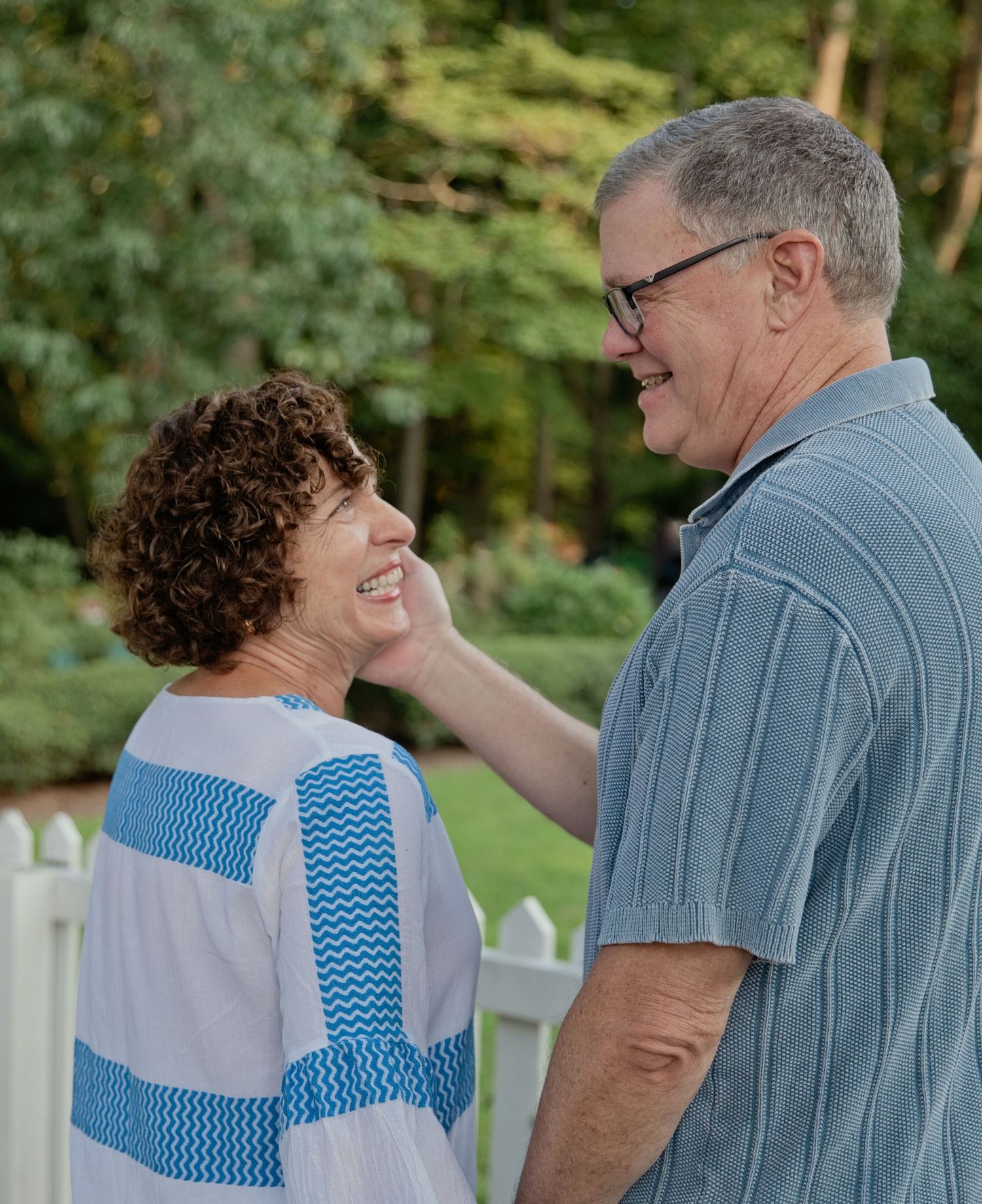 An older couple smiling and sharing a tender moment outdoors near a white fence with green trees behind them.