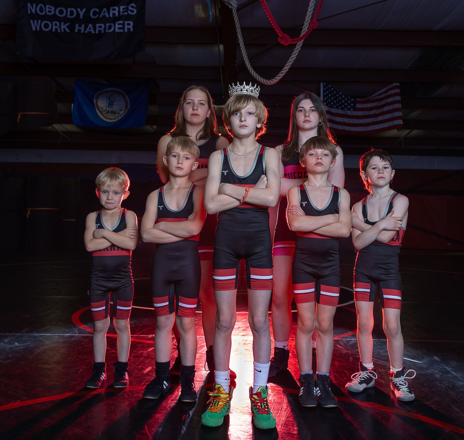 Young wrestlers in black singlets pose confidently in a gym with motivational signage and an American flag.