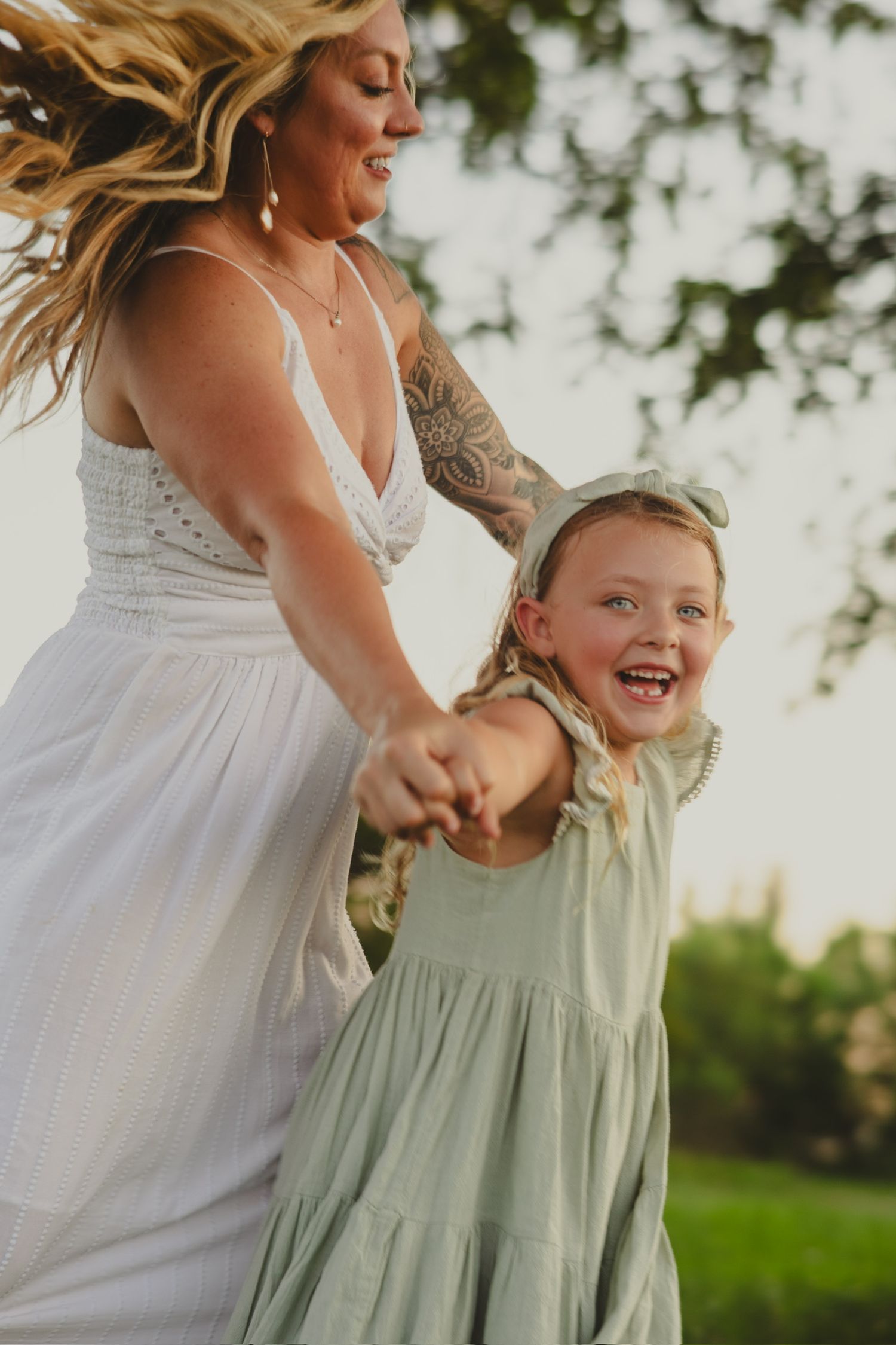 A tattooed woman in white dress spins a laughing little girl in sage green dress outdoors at golden hour.