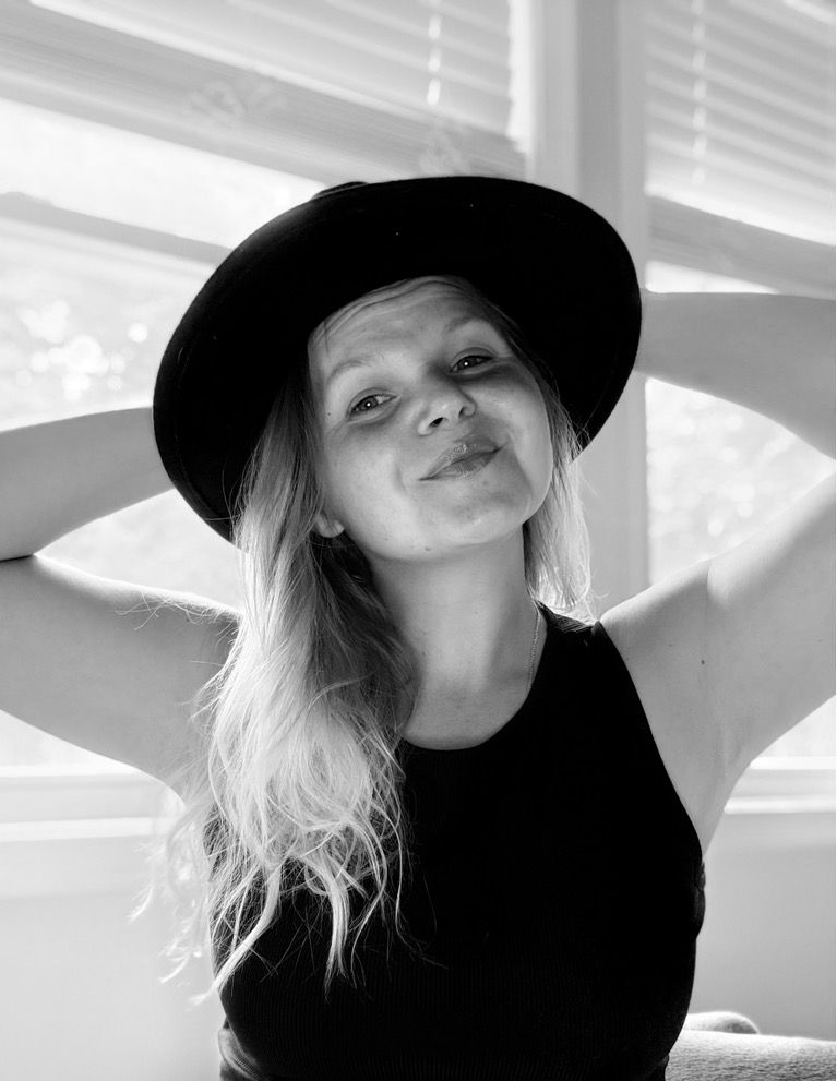 Black and white photo of a young blonde woman wearing a wide-brim black hat, posing with hands behind her head.