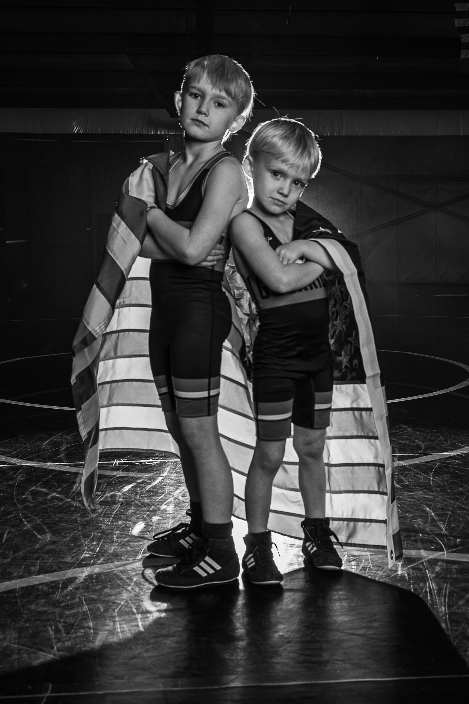 Two young wrestlers in singlets draped in an American flag pose confidently on a mat in a dramatic black and white photo.