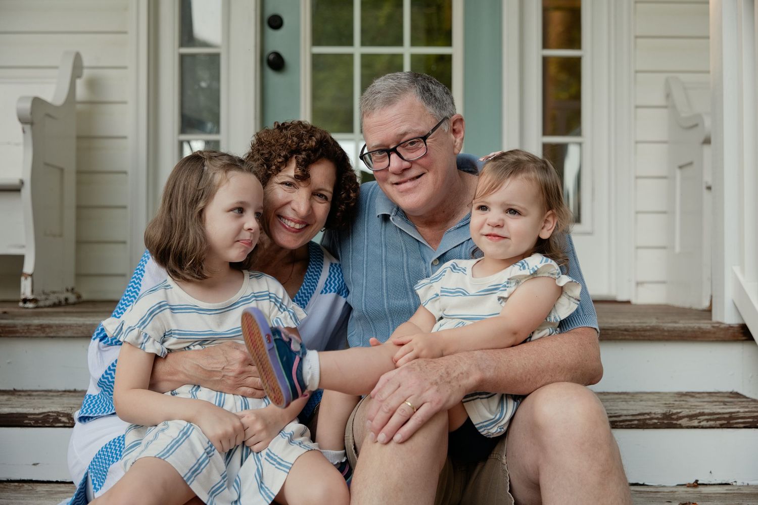 Family of four smiling on porch steps, two young girls in matching dresses with grandparents on a sunny day.