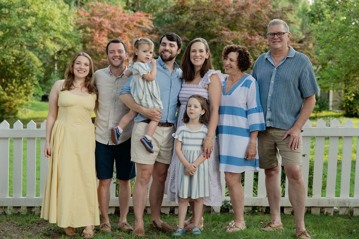 A joyful multigenerational family of eight poses together outdoors near a white picket fence surrounded by lush greenery.