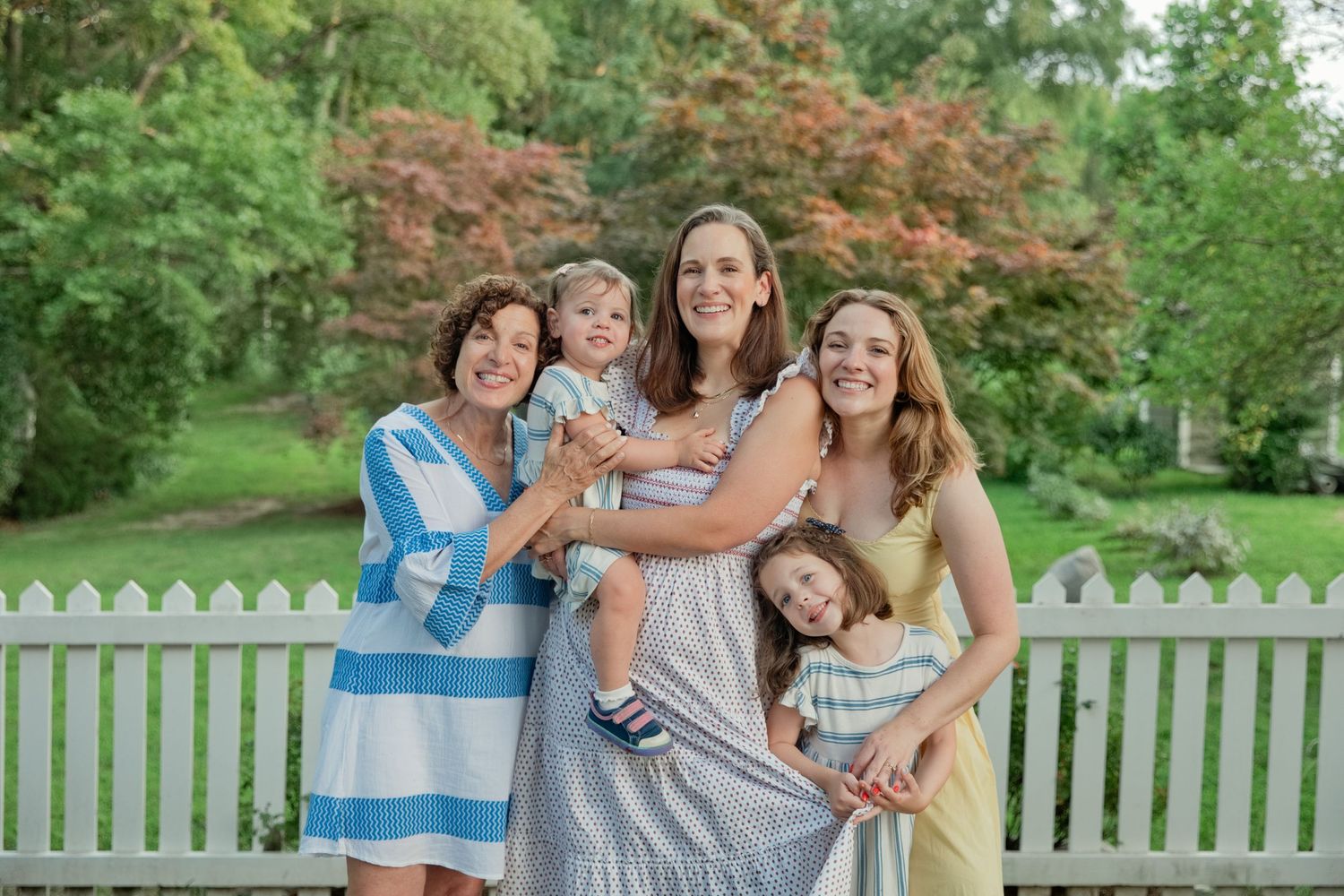A happy multigenerational family of women pose together with two young children by a white fence.